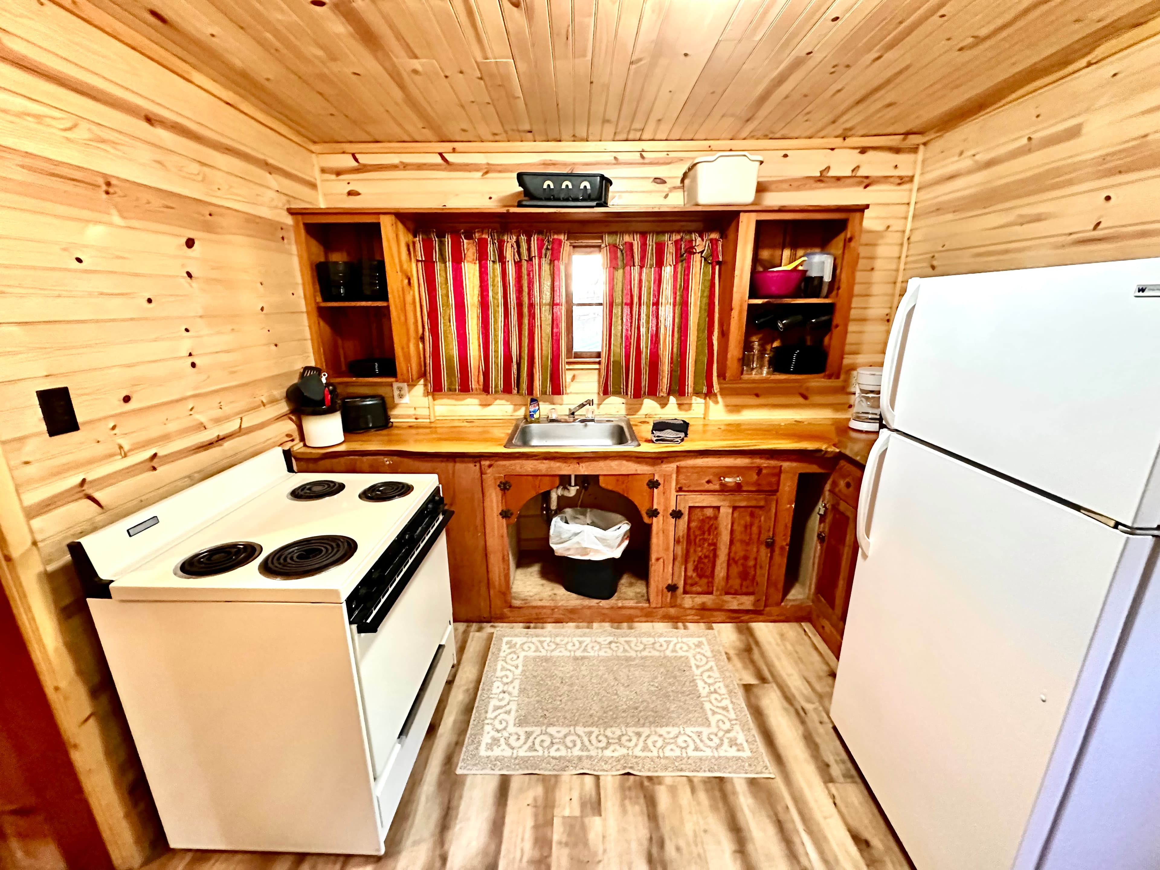 A cozy kitchen with wooden walls, a white stove, a refrigerator, and a sink under a window with red curtains.