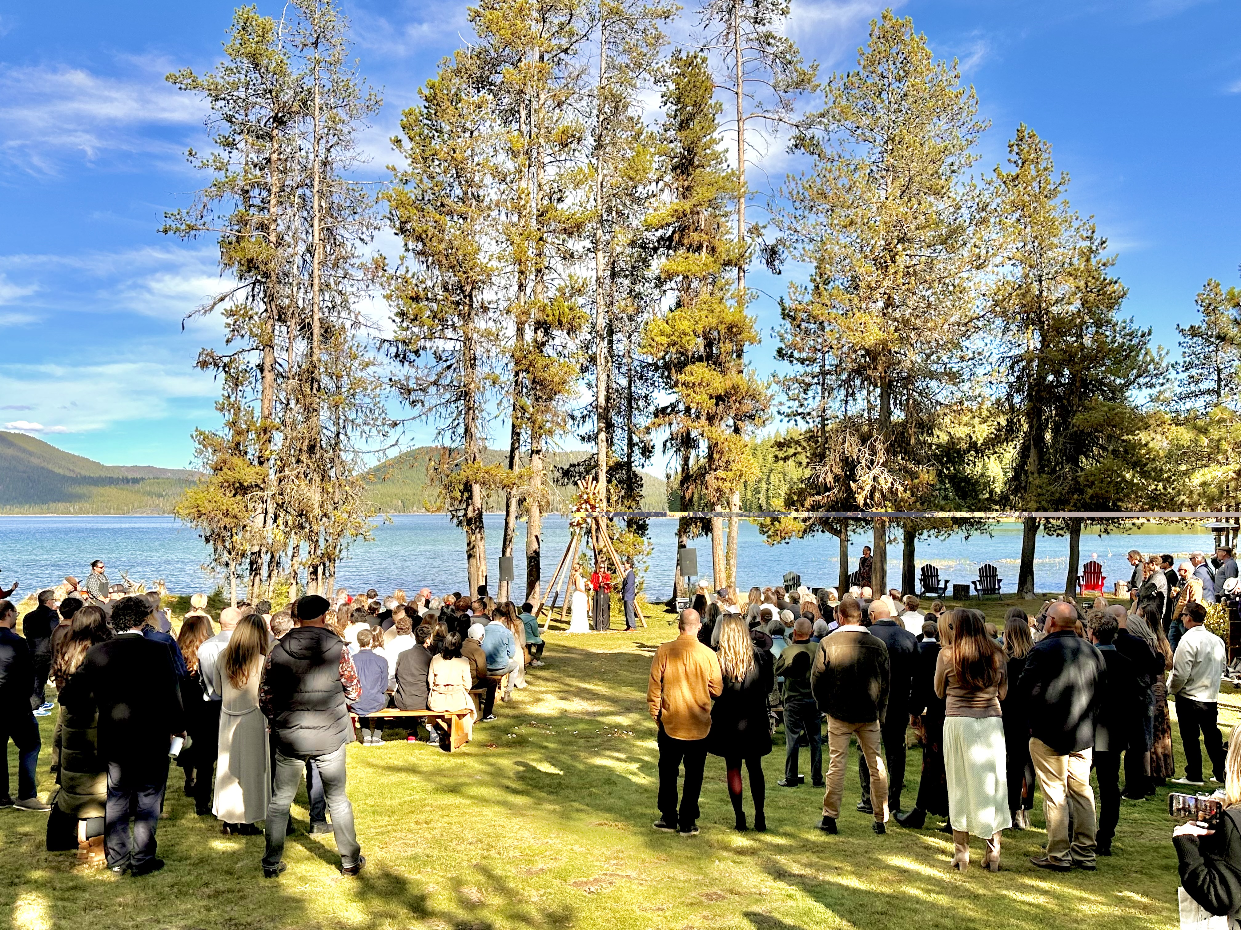 A large outdoor gathering by a lake, where guests are watching a ceremony under tall trees.