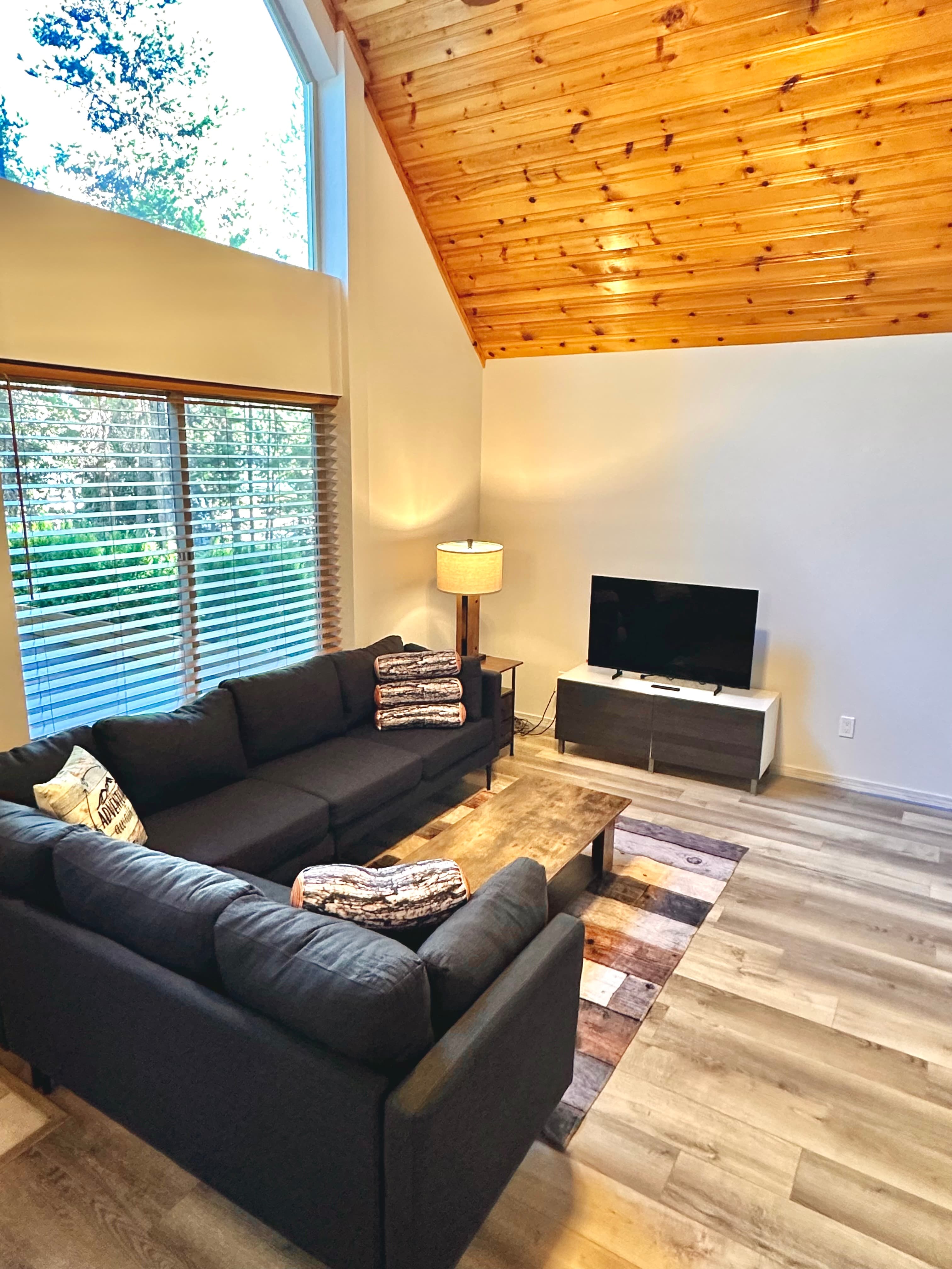 Cozy living room featuring a black sofa, coffee table, TV, and large windows with natural light.