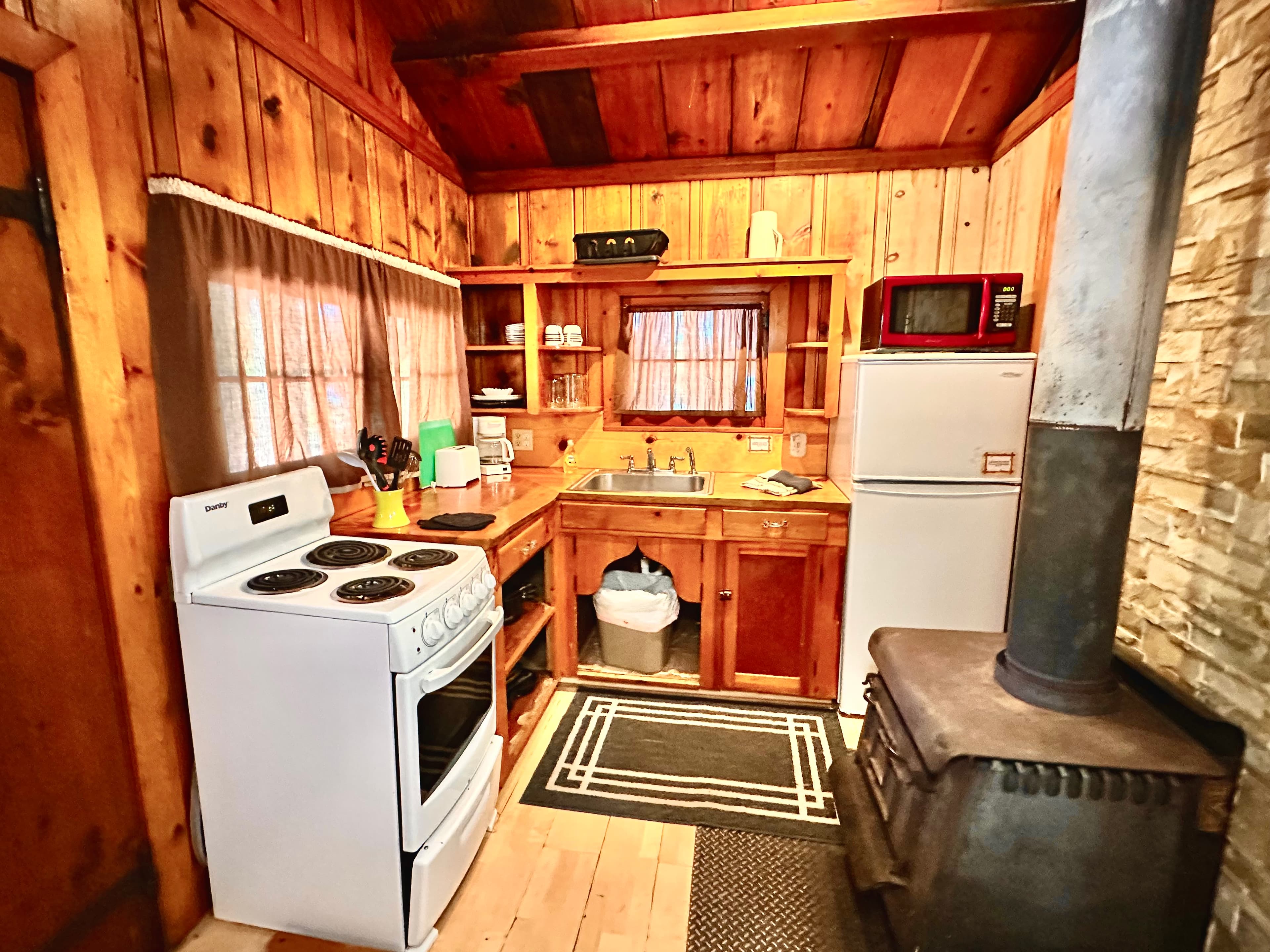 Cozy rustic kitchen featuring wooden walls, a stove, fridge, and stone fireplace.