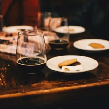 A glass of dark beer beside plates of cookies on a wooden table.