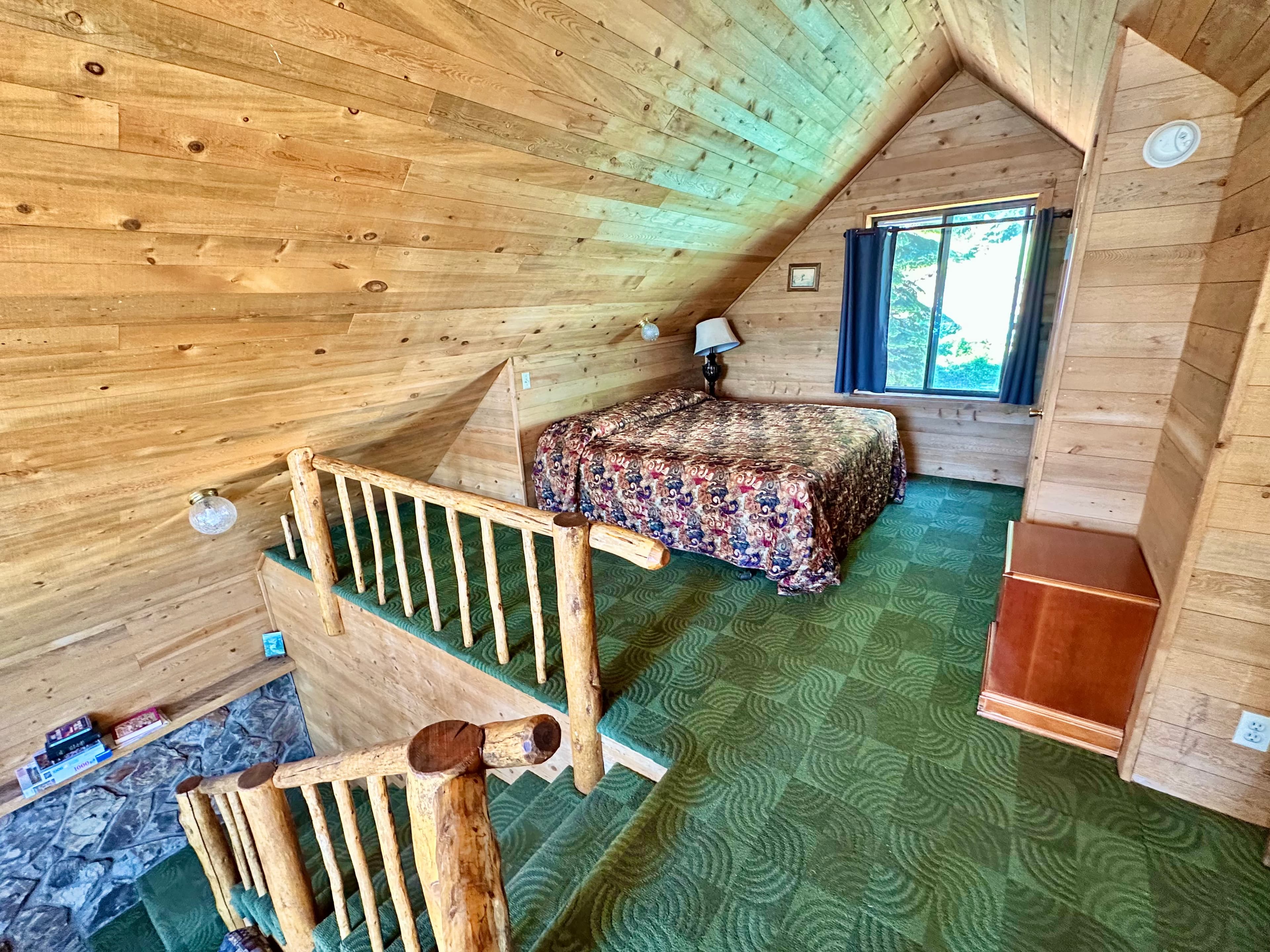 A cozy wooden attic bedroom featuring a patterned bedspread, a window, and a small wooden staircase.