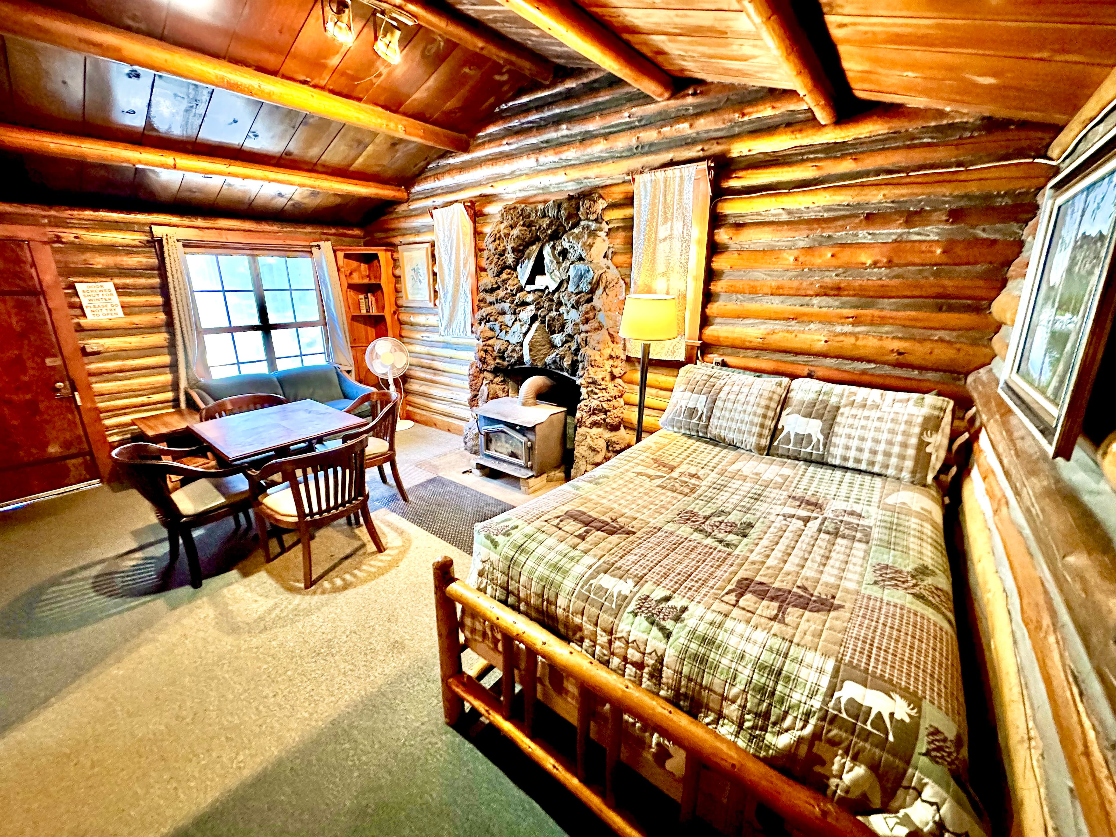 A cozy log cabin interior featuring a bed, dining table, and stone fireplace with rustic decor.