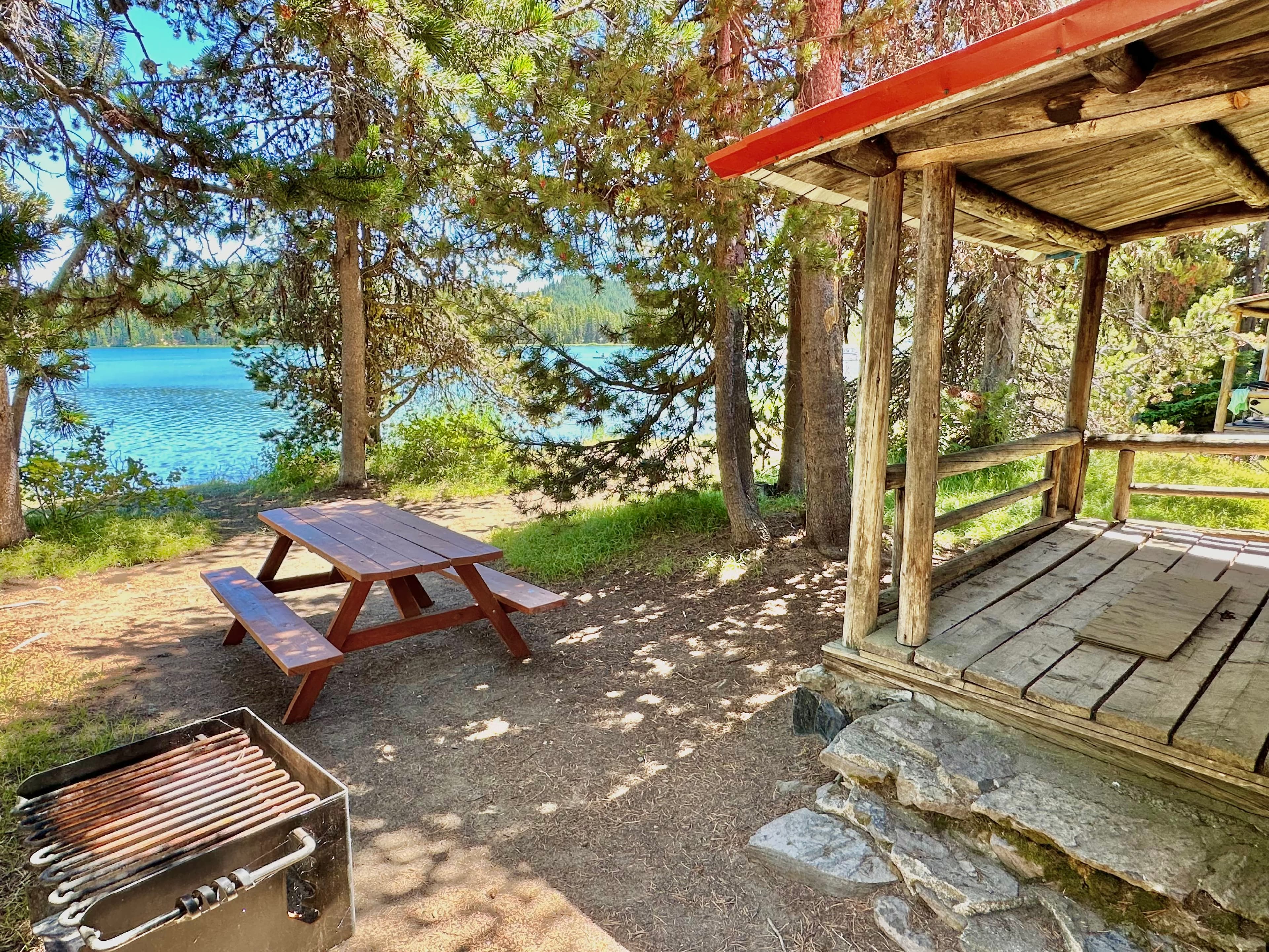 A picnic table and grill beside a serene lake surrounded by trees.