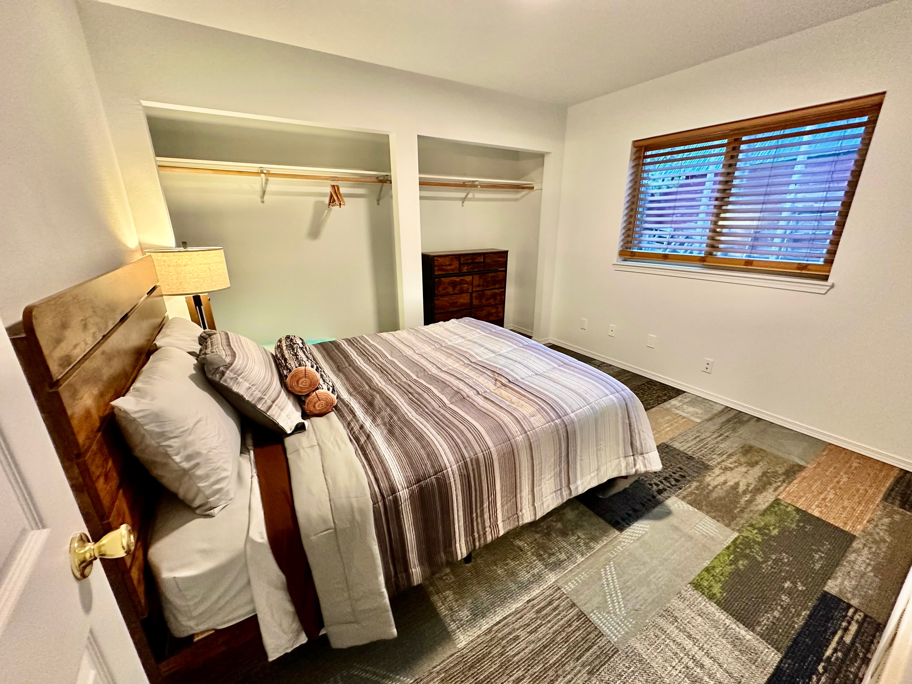 A cozy bedroom featuring a striped bedspread, a wooden bed frame, a lamp, and a window with blinds.