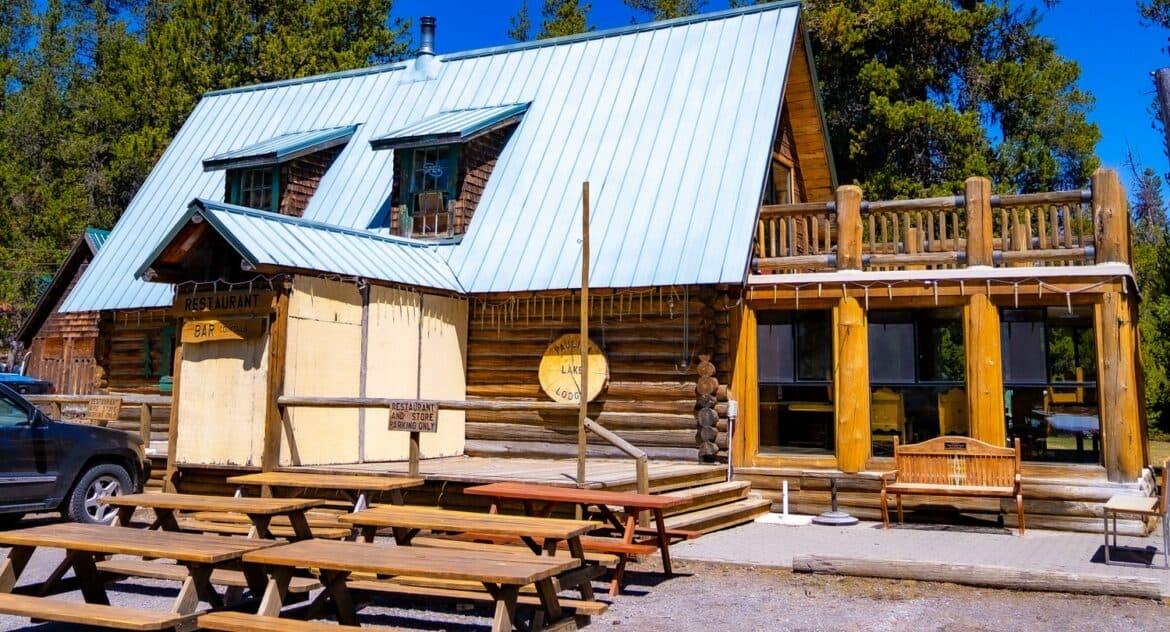 A rustic log cabin with a metal roof, featuring picnic tables in front and a porch with seating.