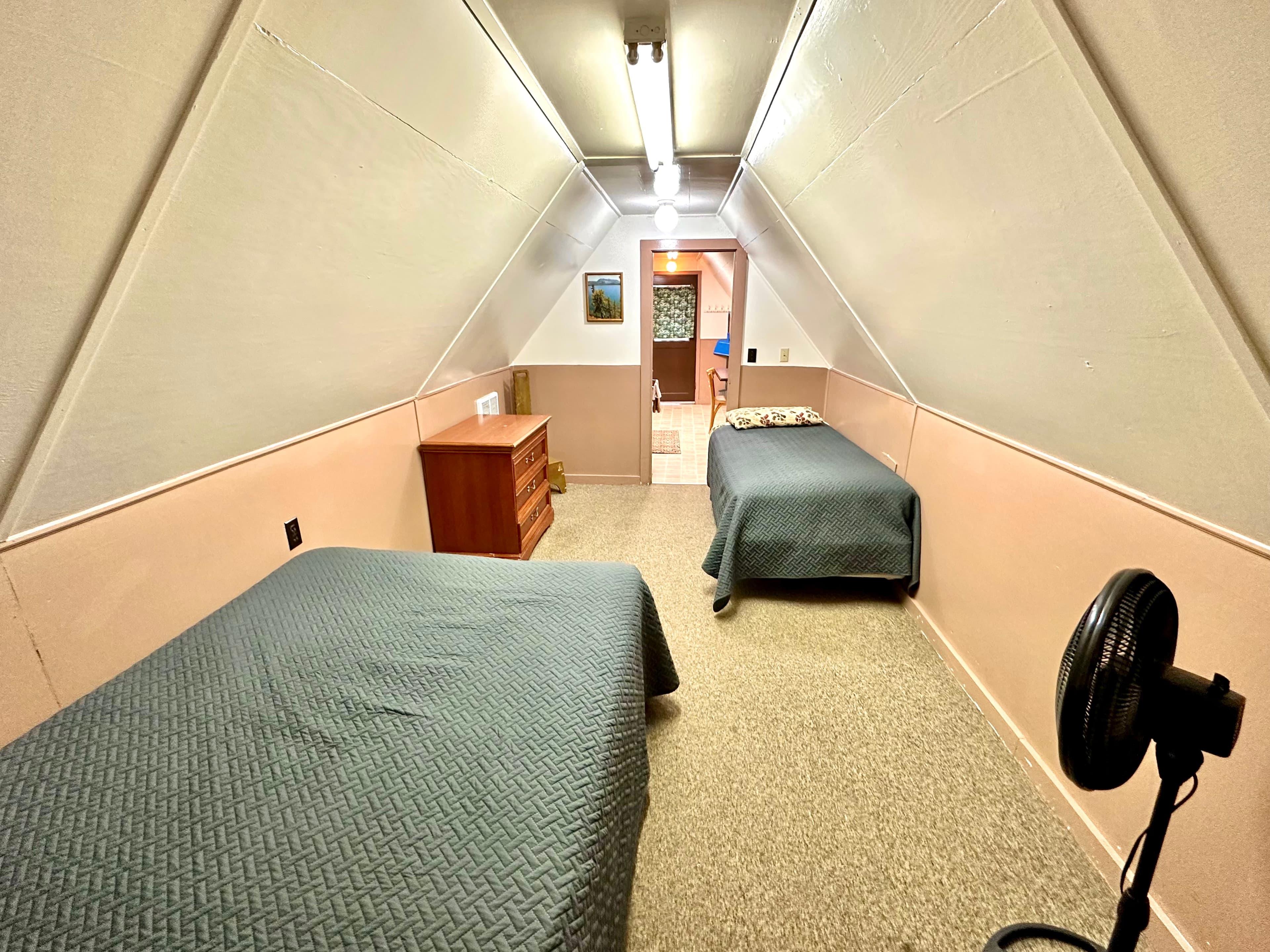 A narrow, triangular-shaped room featuring two twin beds, a wooden dresser, and a fan.