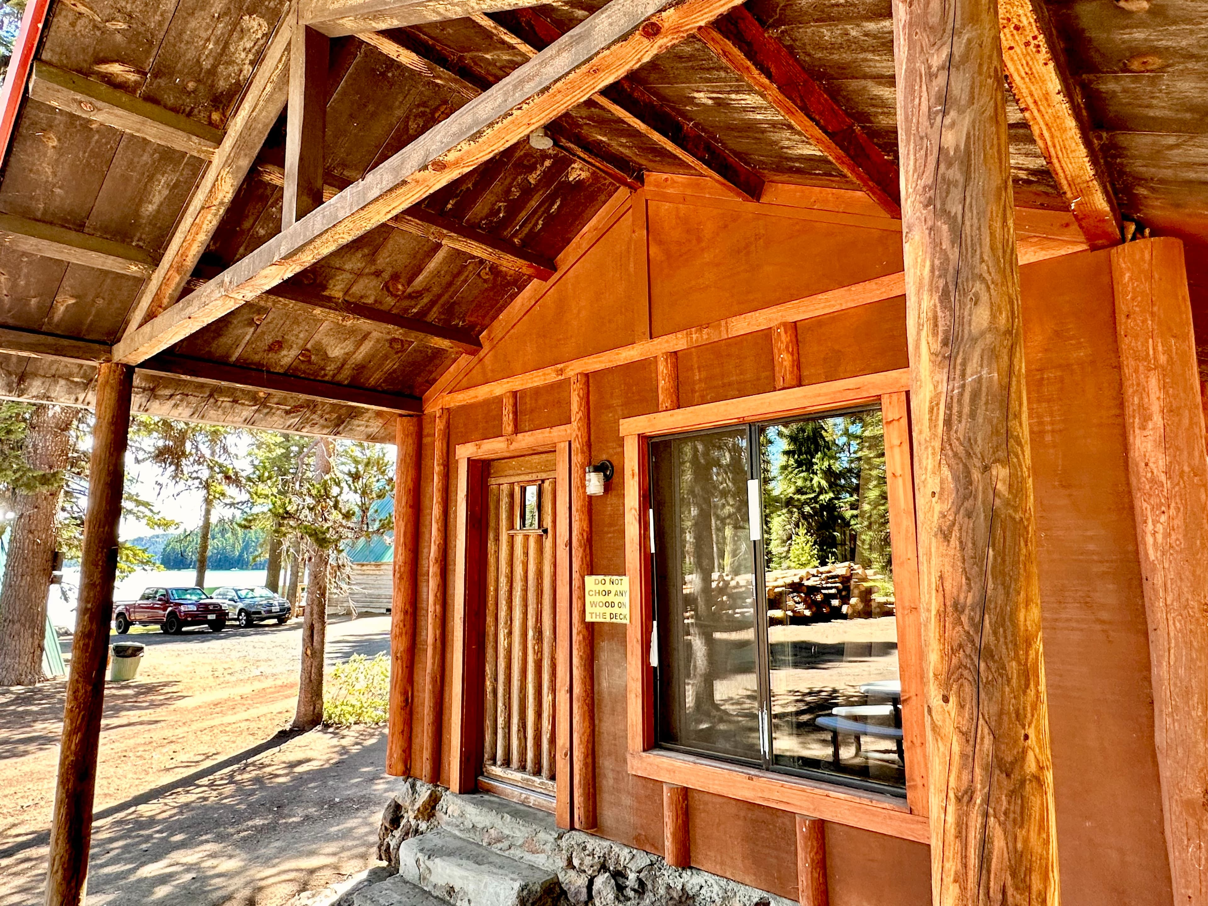A rustic cabin with wooden beams and a large window, set against a backdrop of trees and parked cars.