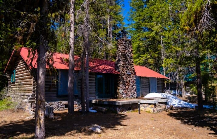 A rustic log cabin with a red roof and stone chimney nestled among tall trees.