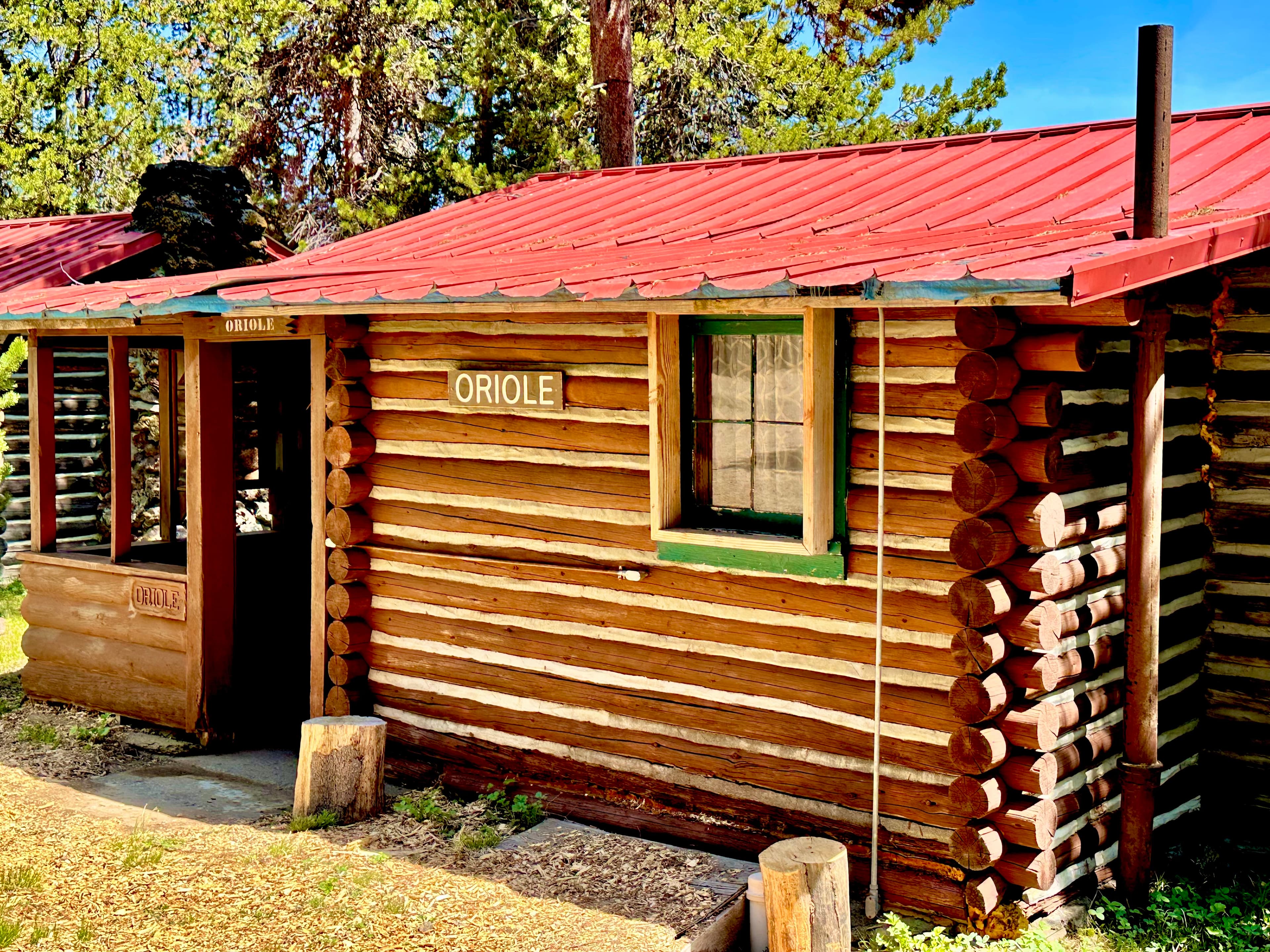A rustic log cabin with a red metal roof, labeled "ORIOLE," surrounded by trees.