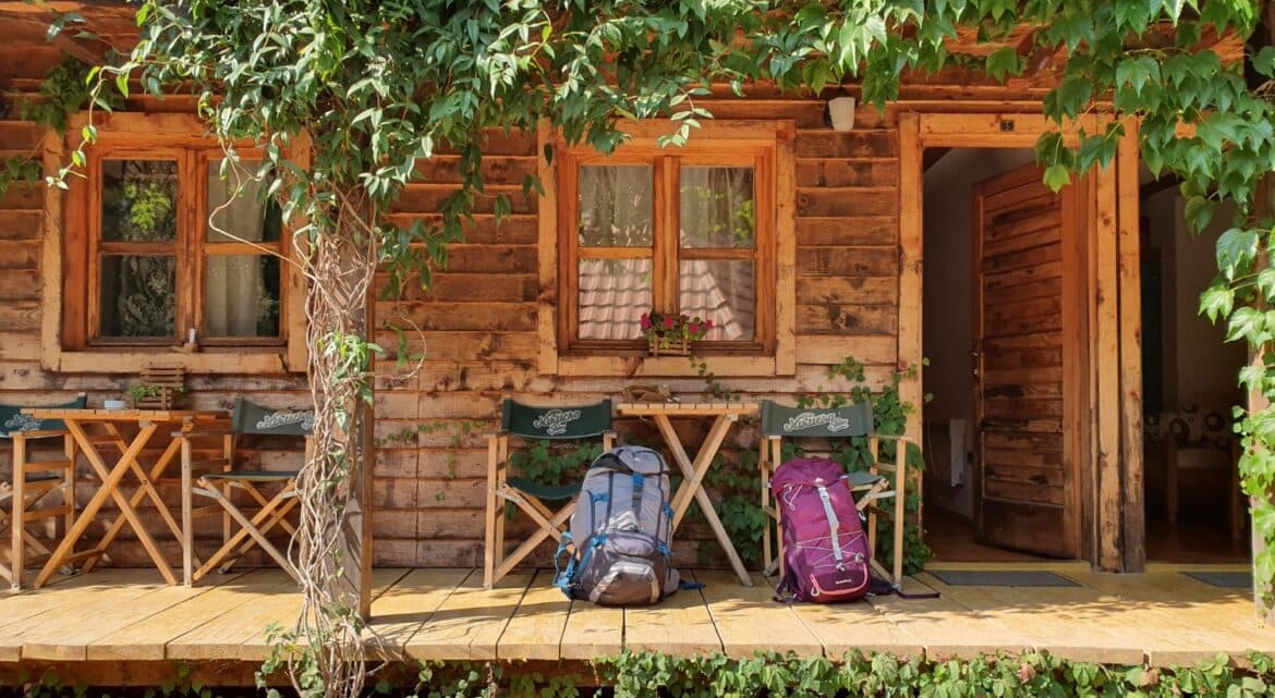 A rustic wooden cabin porch with two backpacks and folding chairs surrounded by greenery.