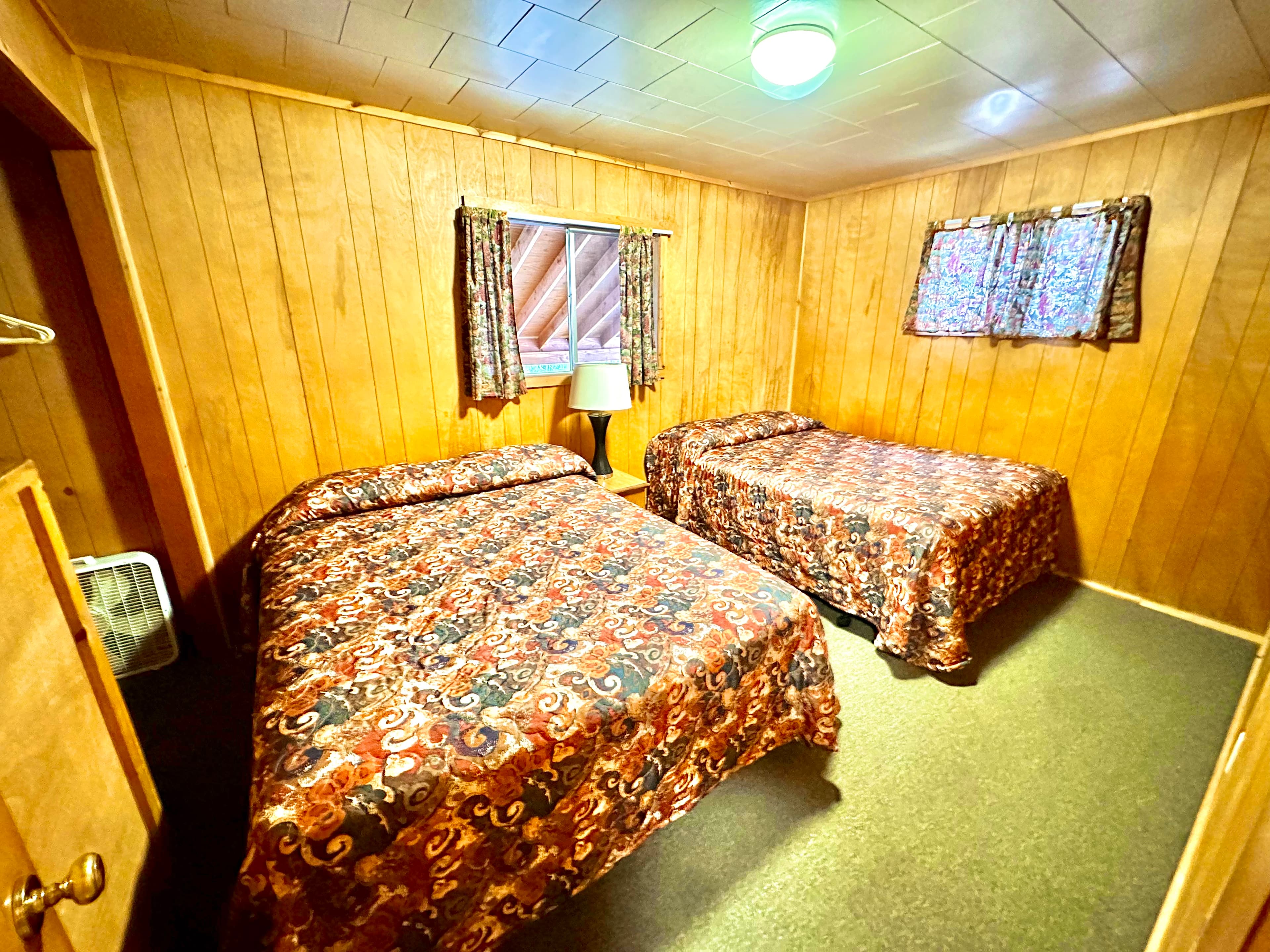A cozy room featuring two beds with patterned blankets, wooden paneling, and a window with curtained natural light.