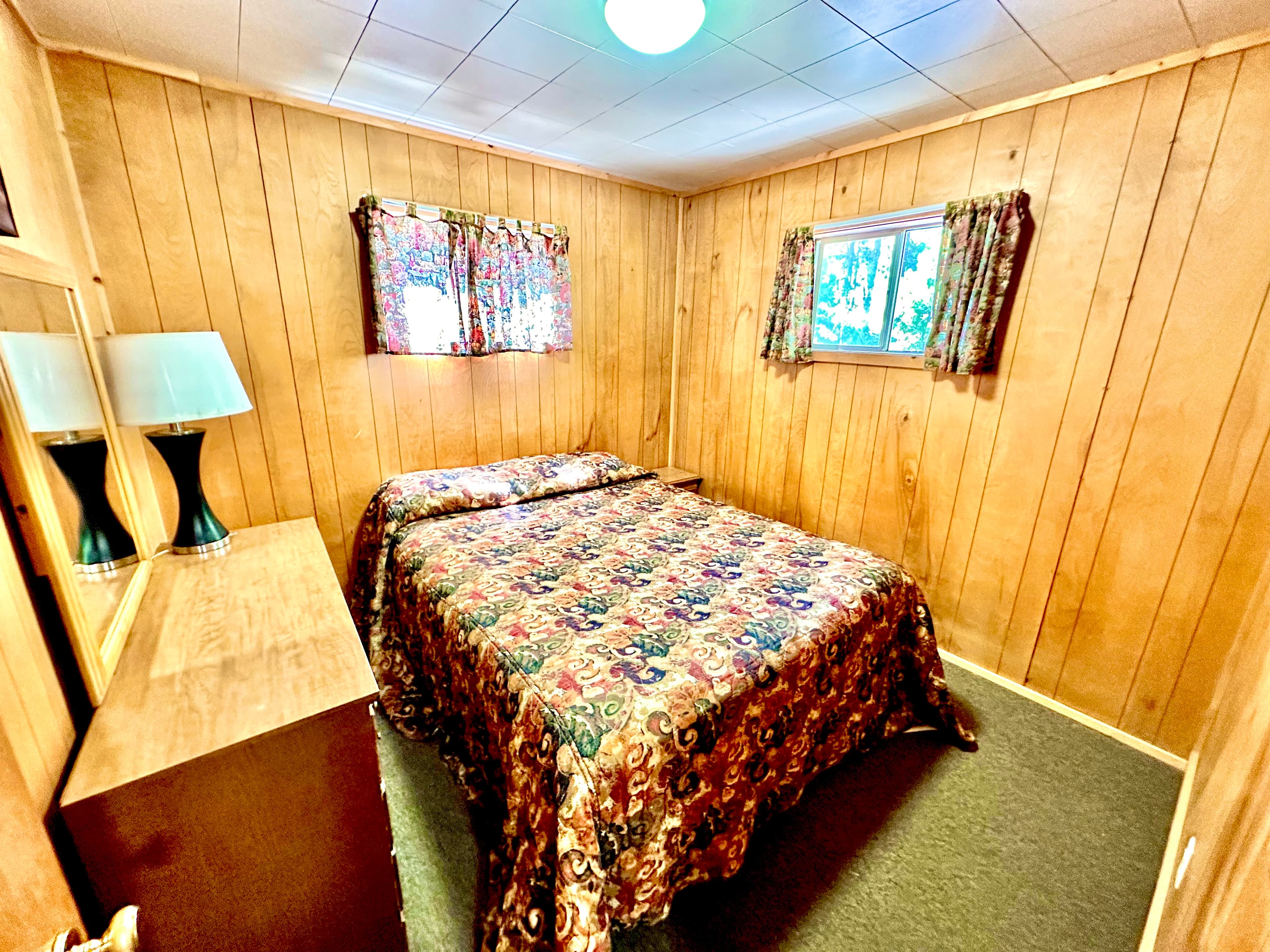 A cozy bedroom featuring a double bed with a floral bedspread, wooden paneling, and a lamp on a bedside table.