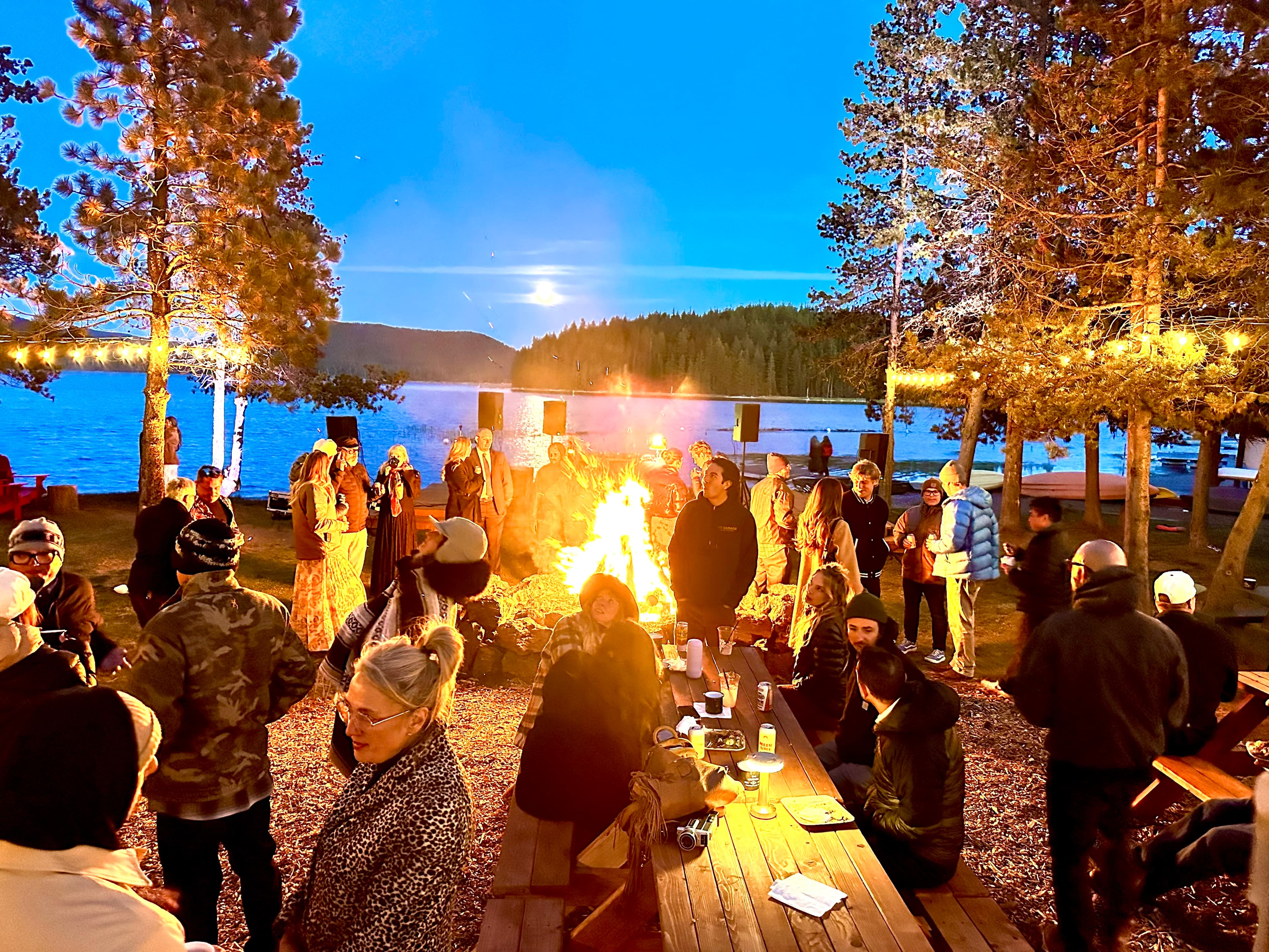 A lively gathering of people around a bonfire by a lakeside at twilight.