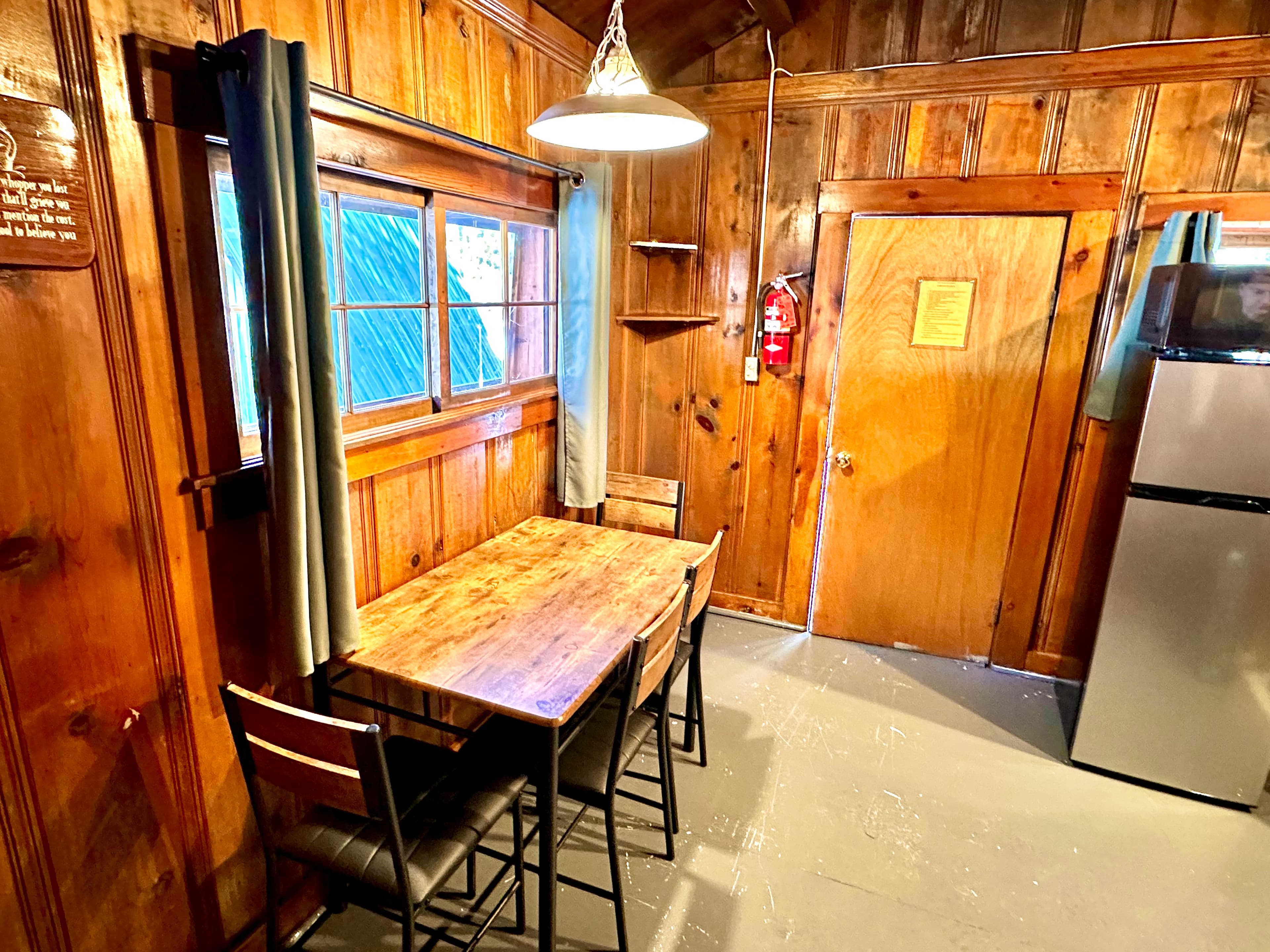 A small wooden dining area with a table and chairs, featuring a window and an entrance door in a cozy cabin setting.
