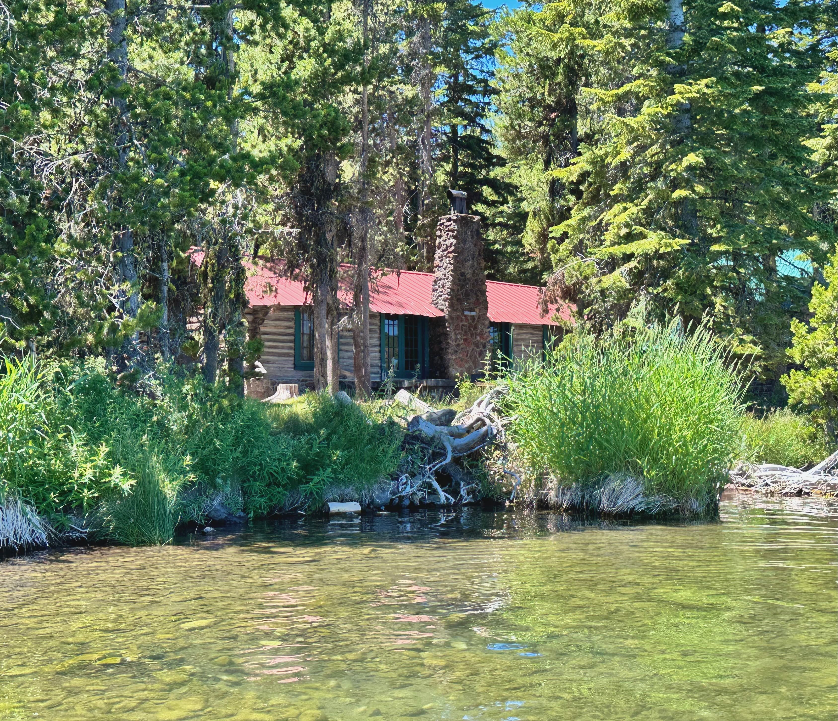 A rustic cabin with a red roof is surrounded by lush green trees and grasses, viewed from a calm lake.