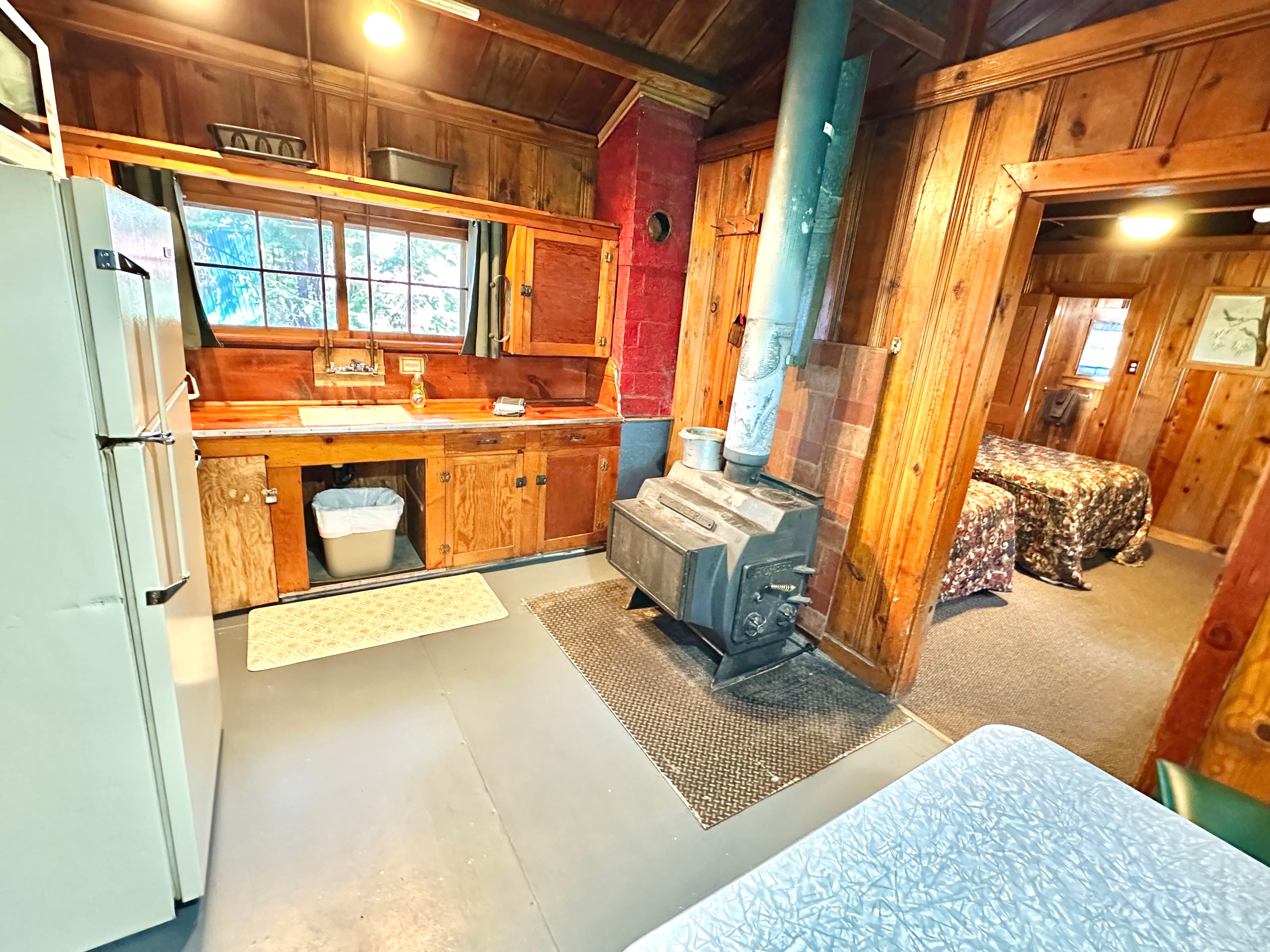 A cozy cabin interior featuring a kitchen area, wood stove, and adjoining bedroom.