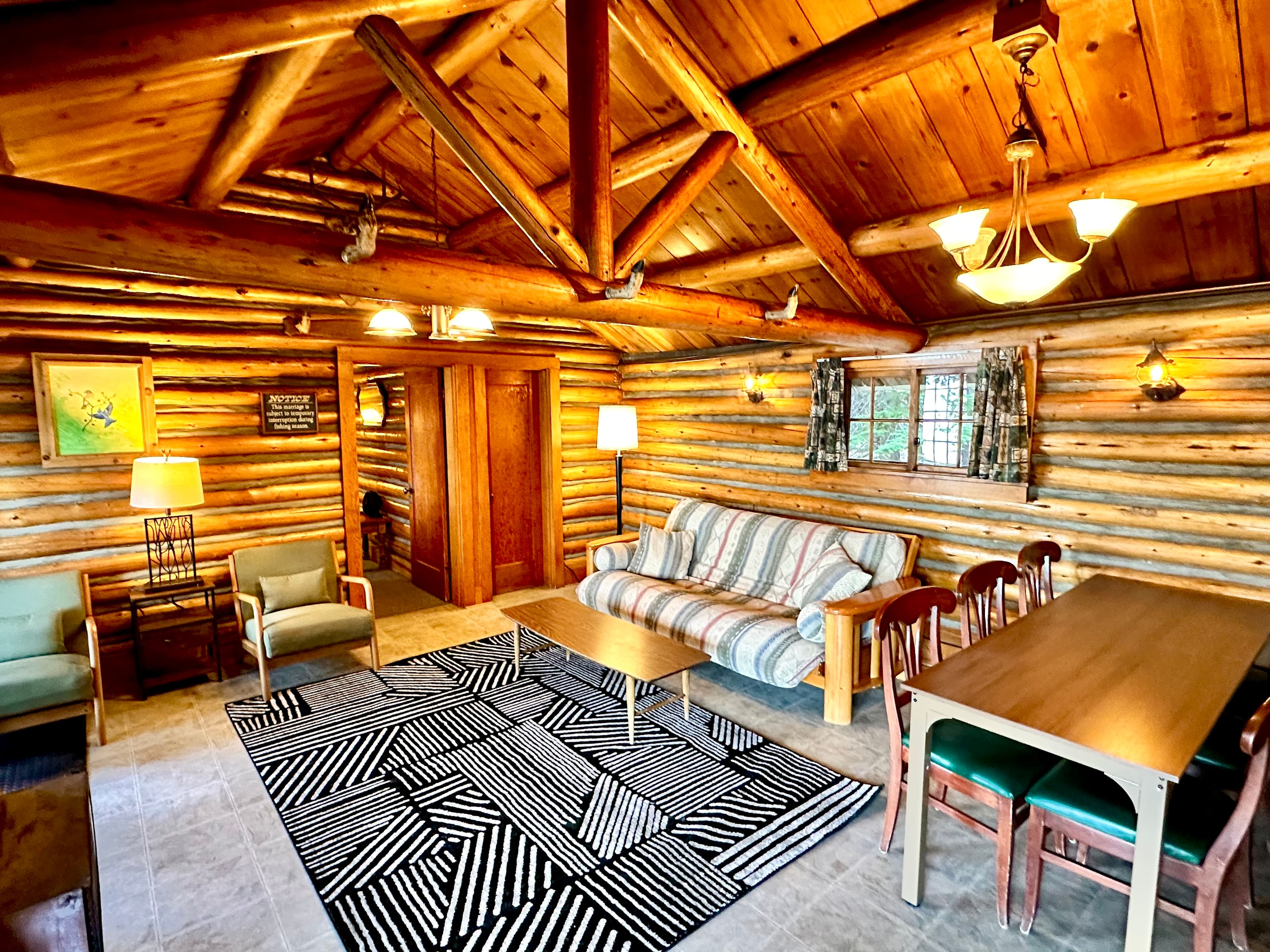 A cozy log cabin interior with wooden beams, a striped rug, seating areas, and a dining table.