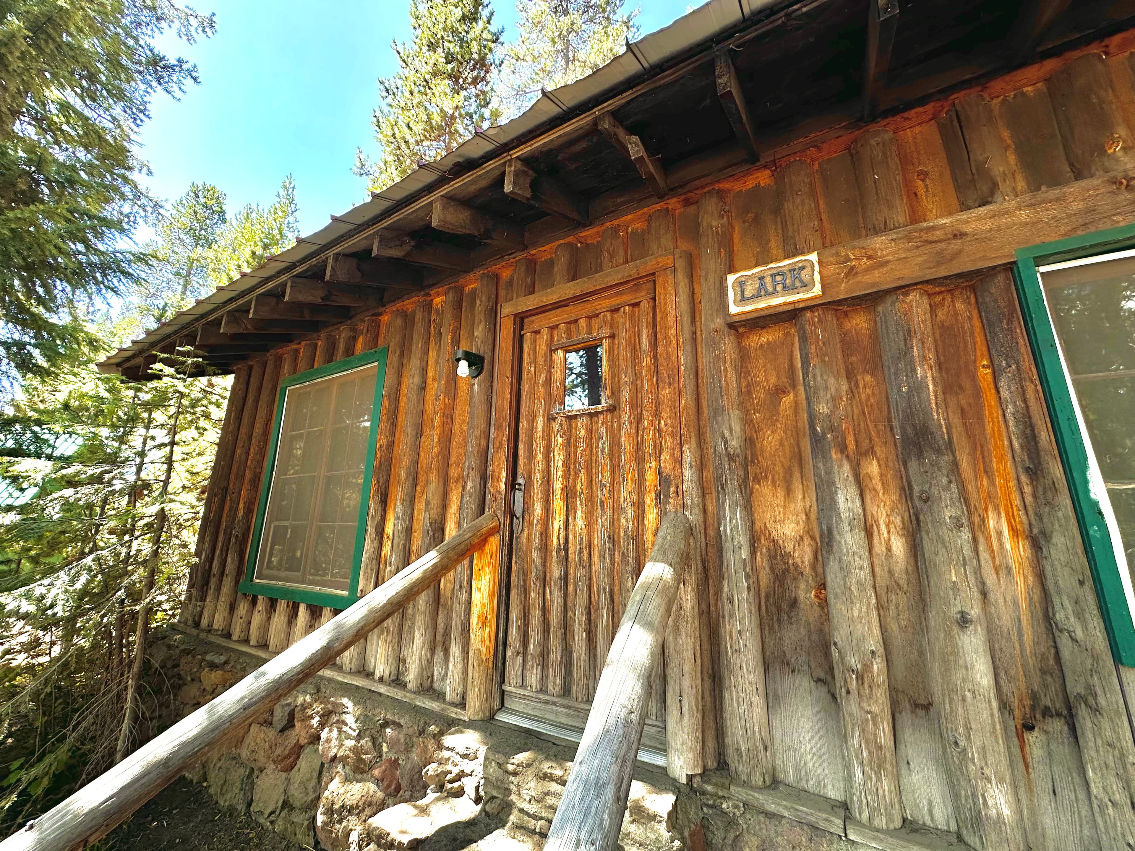 A rustic wooden cabin with a green-trimmed door and sign labeled "LARK," surrounded by trees.