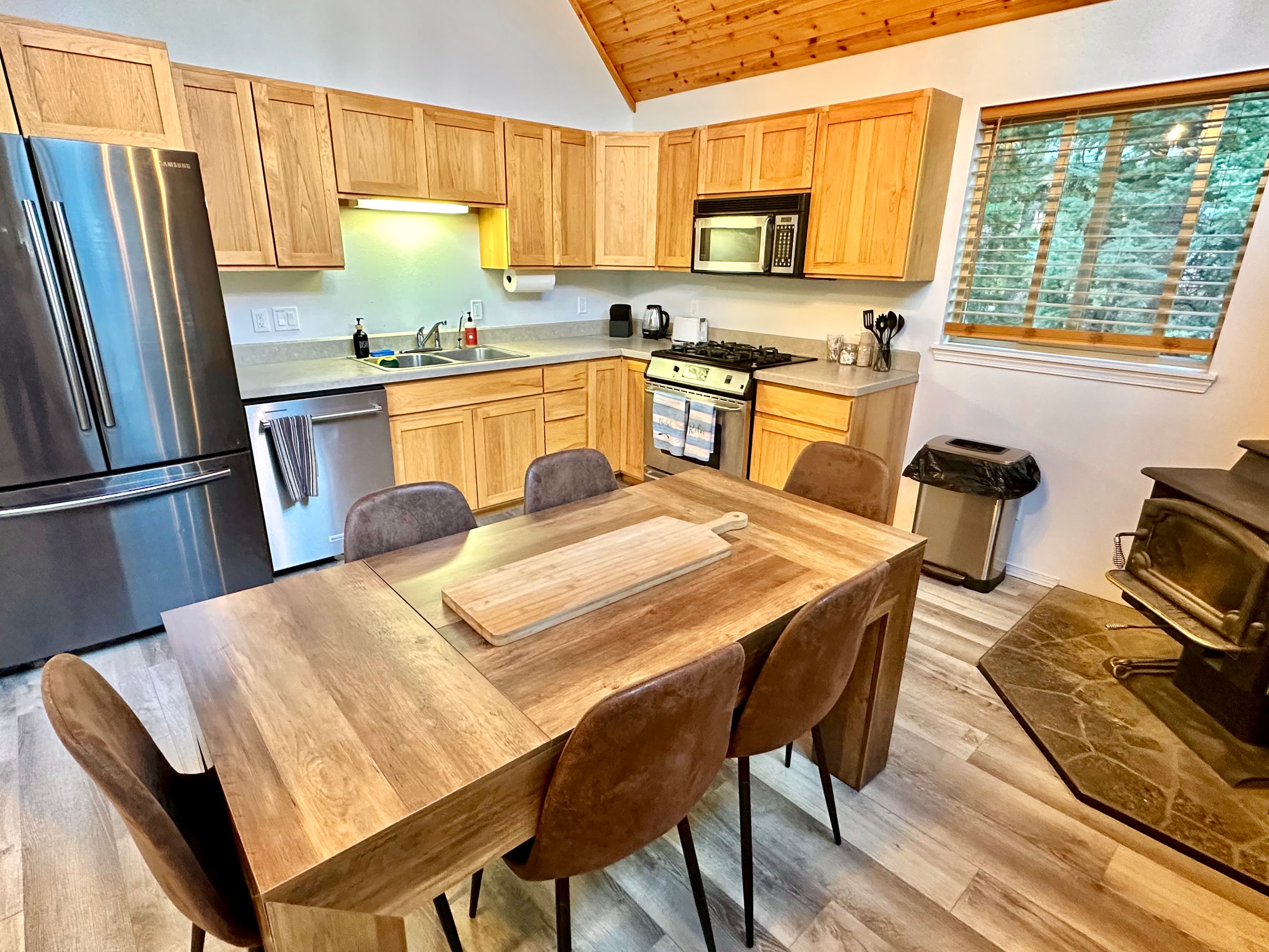 A modern kitchen featuring wooden cabinets, stainless steel appliances, a dining table with chairs, and a nearby wood stove.