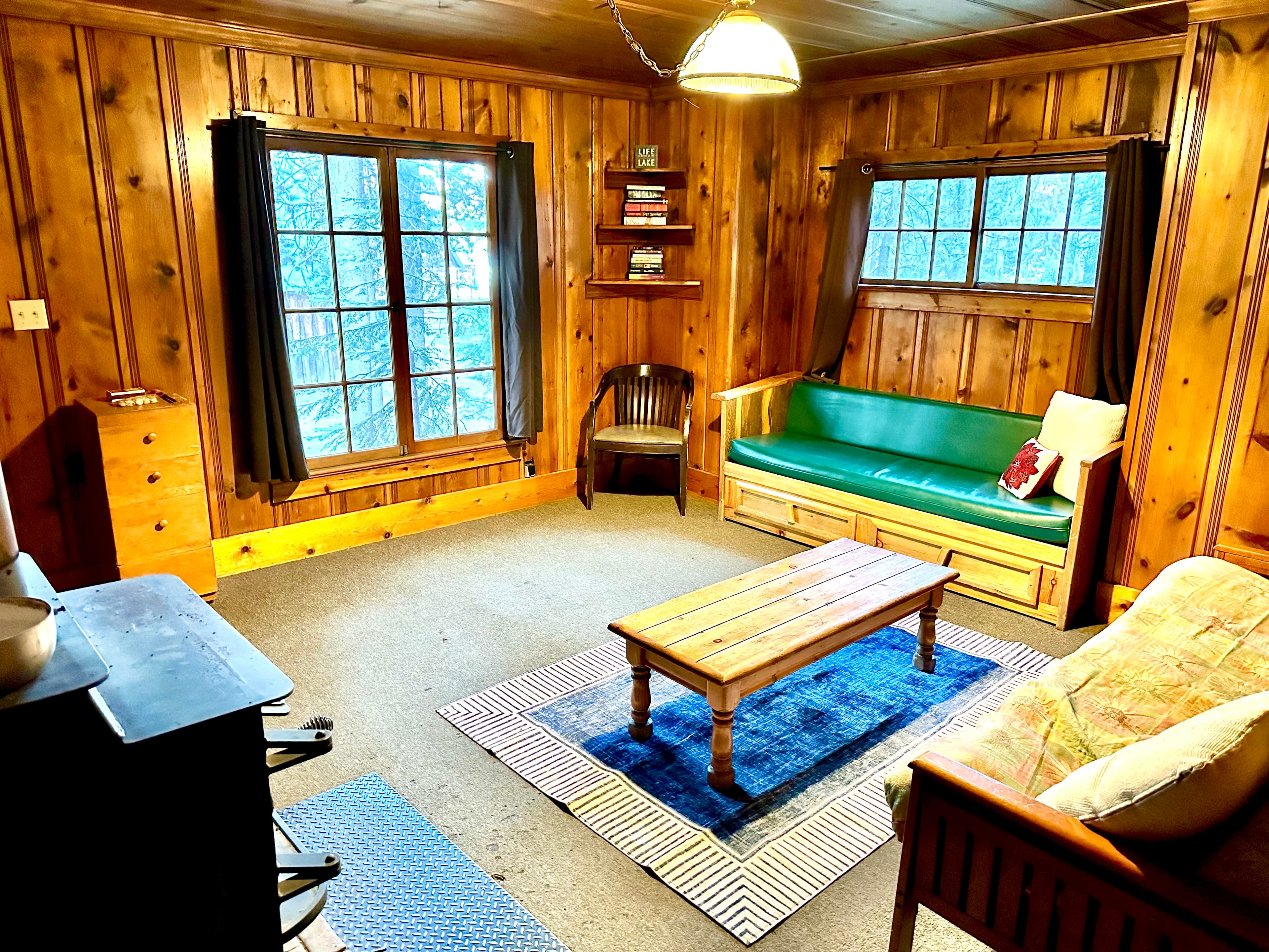 A cozy wooden cabin living room featuring two windows, a green sofa, a wooden coffee table, and bookshelves.