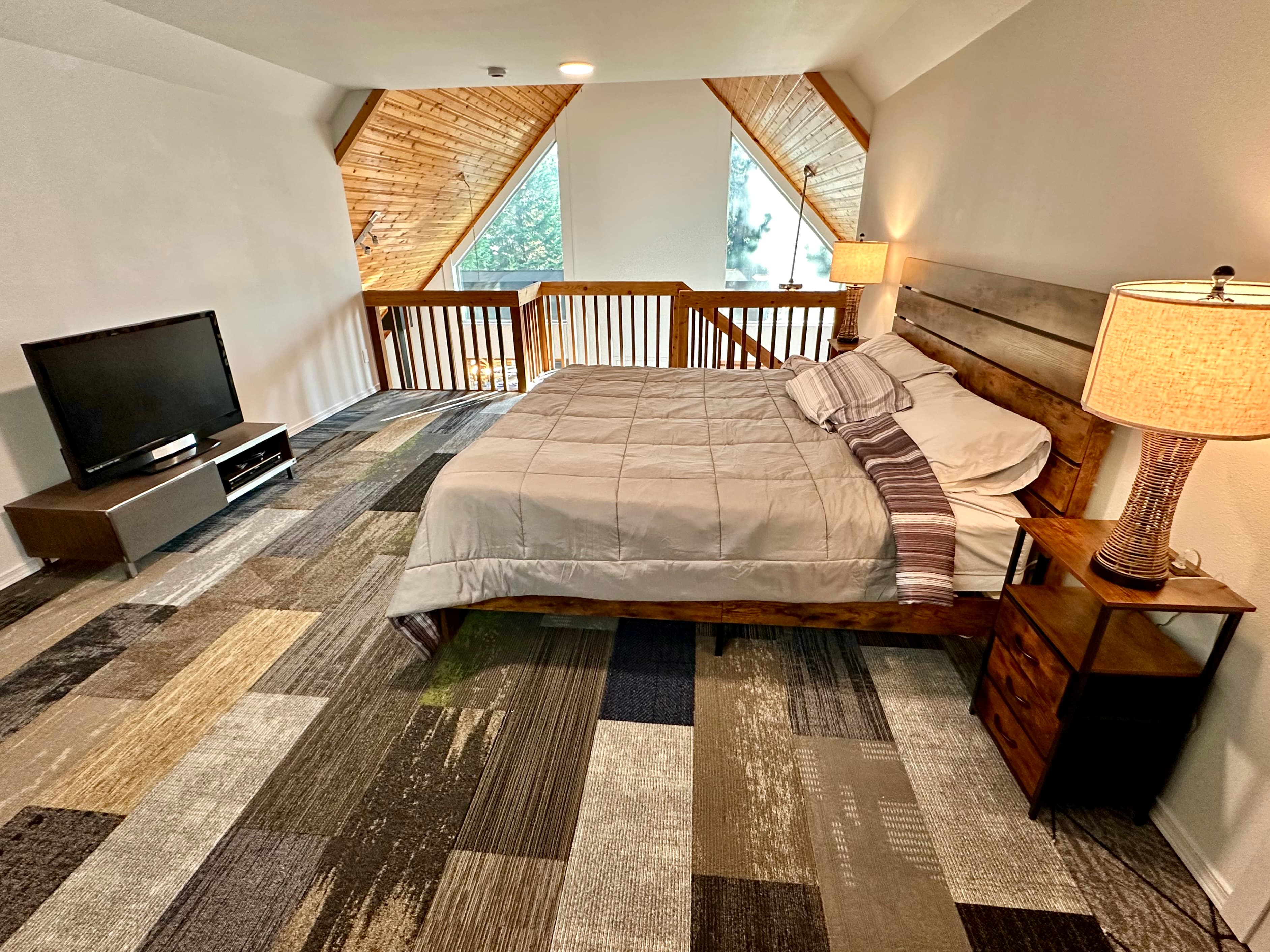 A cozy bedroom with a wooden bed, TV, and a lamp, featuring large windows and sloped ceilings.