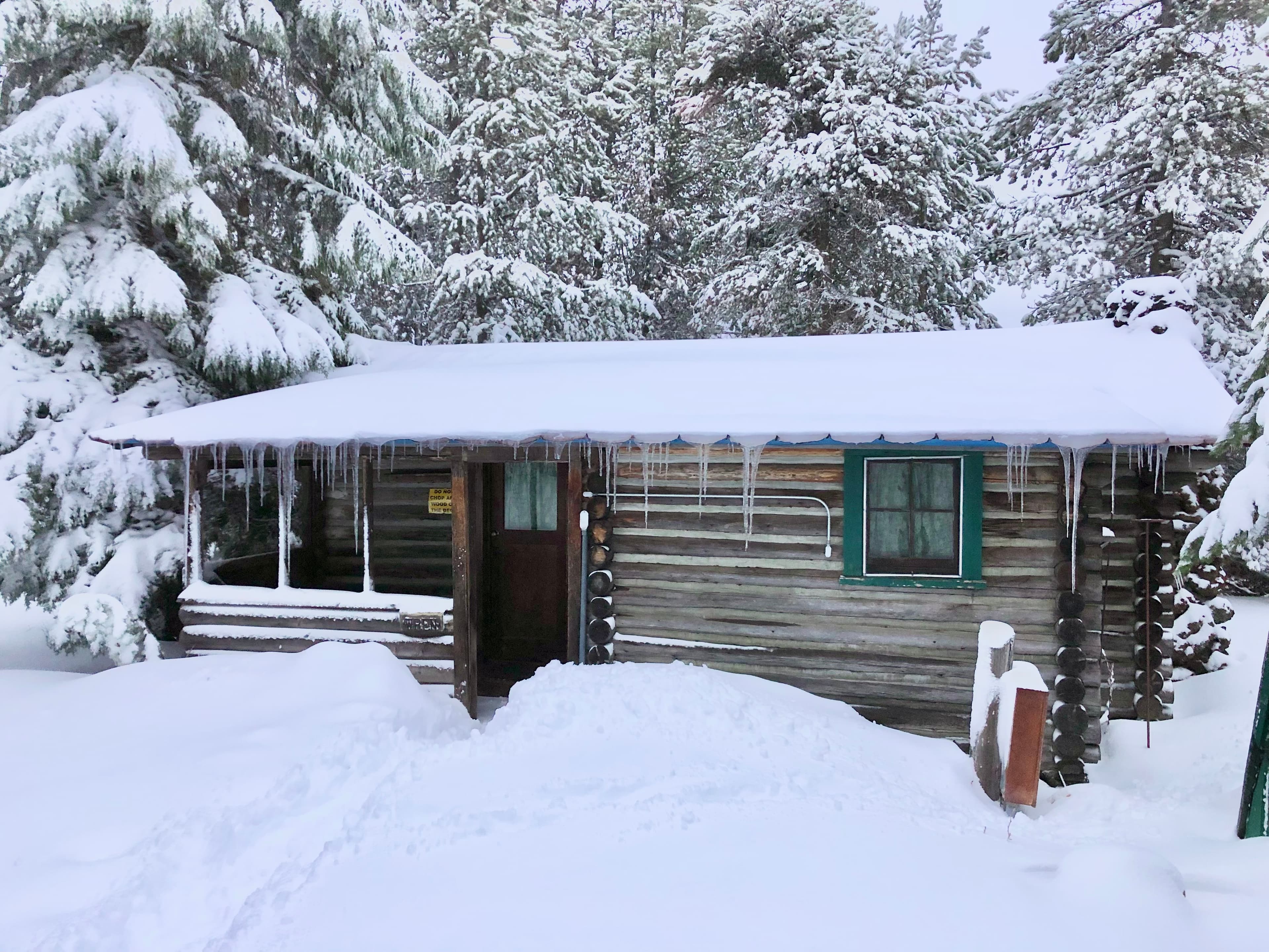 A rustic log cabin surrounded by deep snow and icicles hanging from the roof.