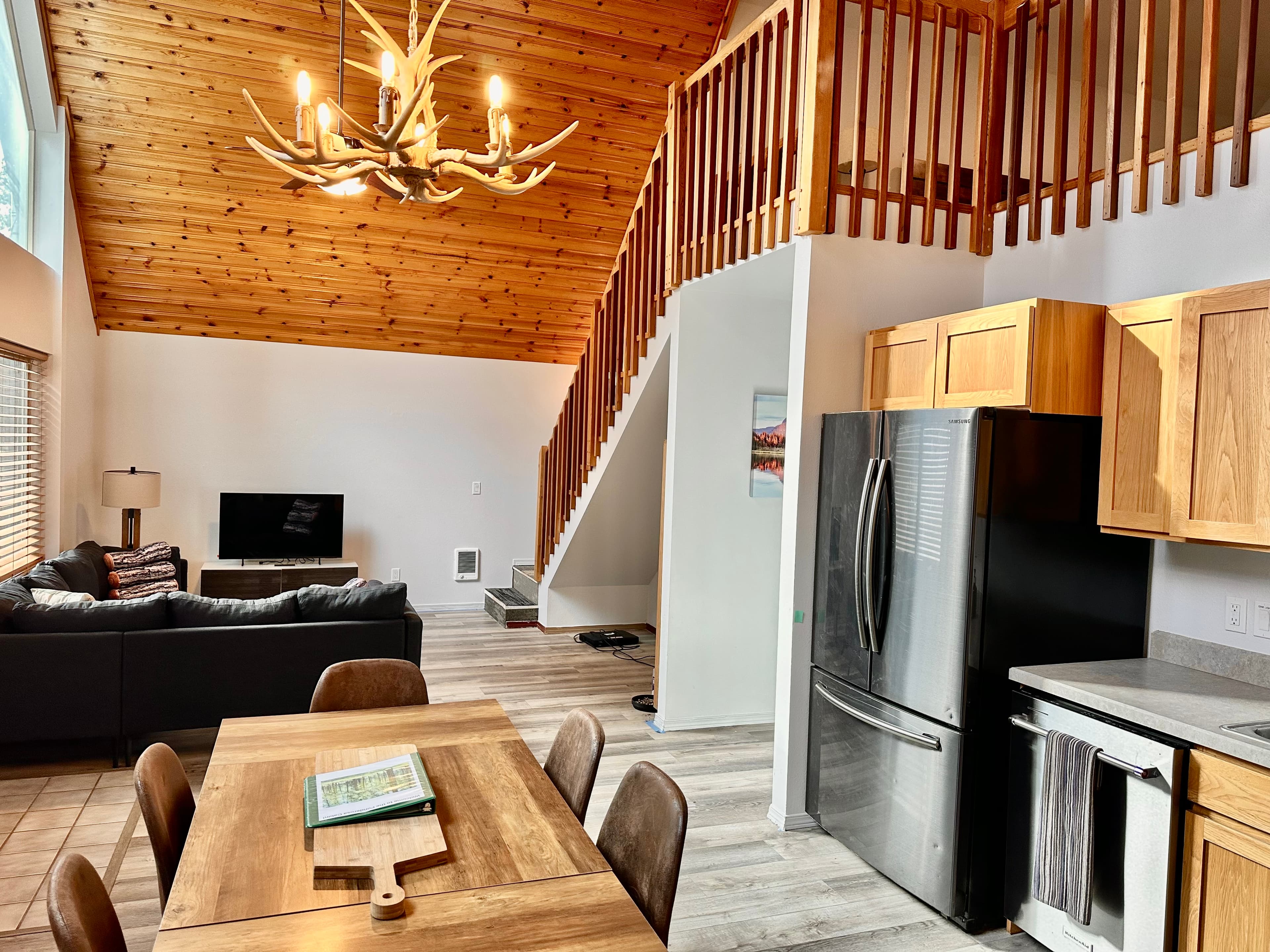A cozy kitchen and living room with wooden accents, featuring a dining table, a black sofa, and a modern refrigerator under a high, vaulted ceiling.