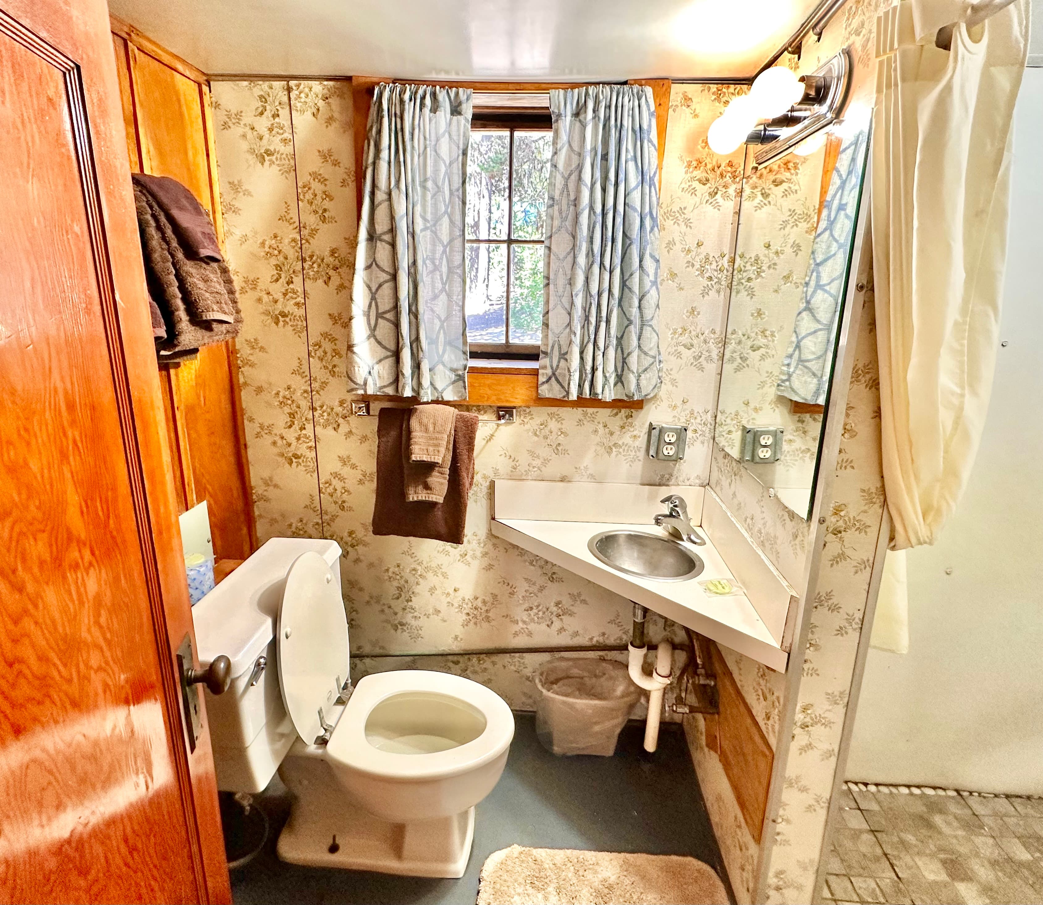 A small, rustic bathroom featuring a toilet, sink, window with sheer curtains, and floral wallpaper.