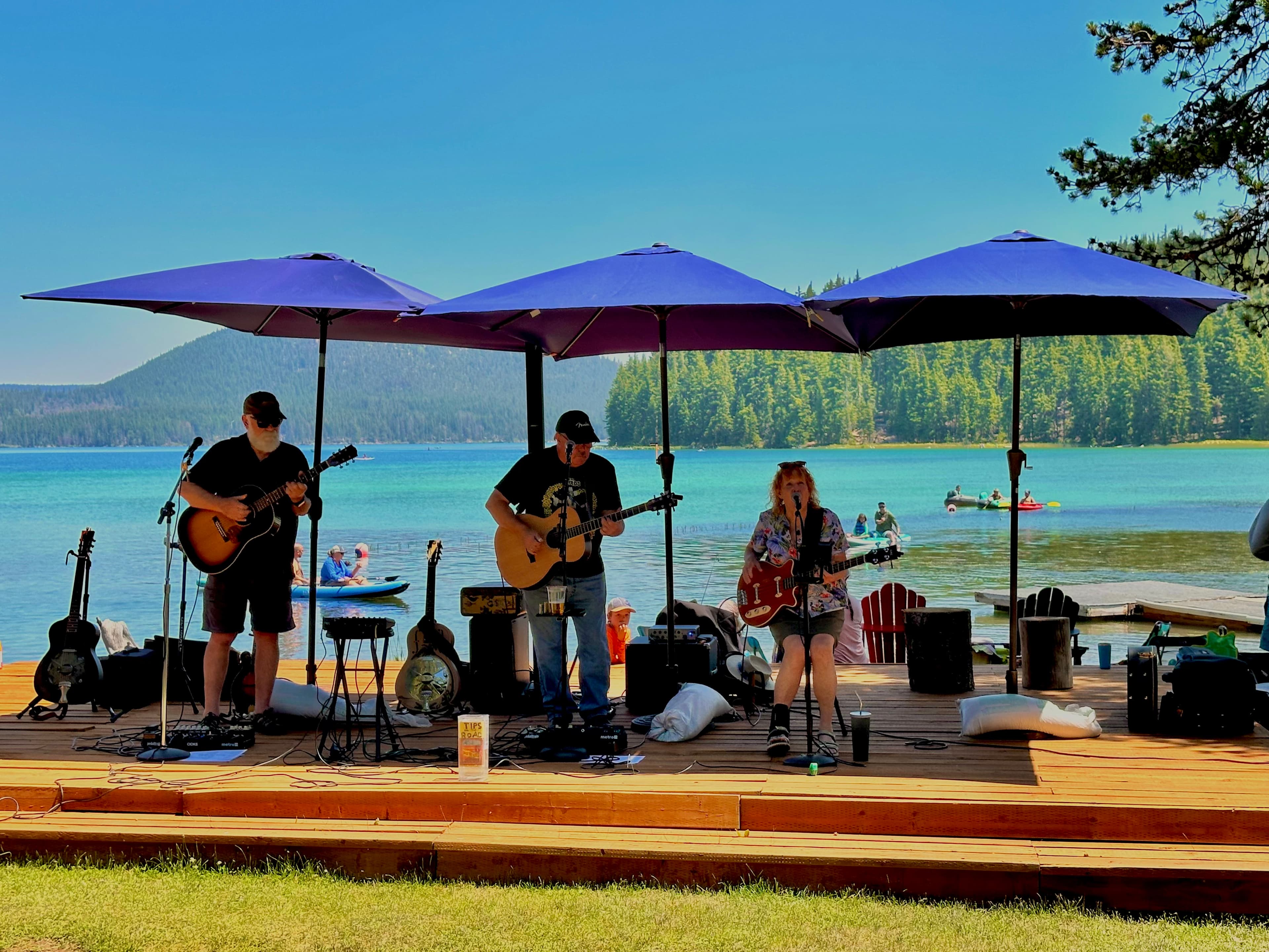 Three musicians perform on a lakeside stage with blue umbrellas, surrounded by kayakers and a scenic landscape.