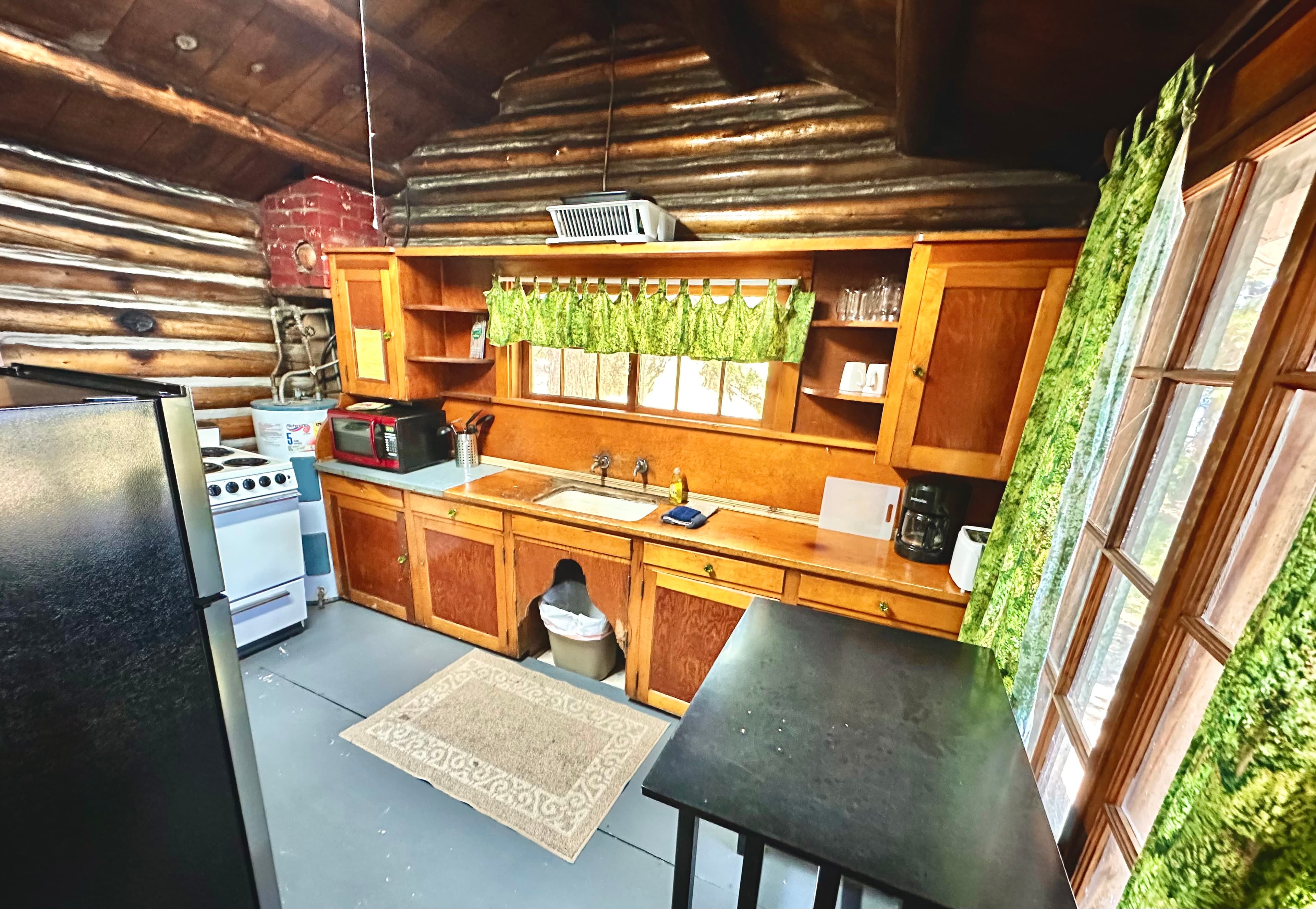 A rustic cabin kitchen featuring wooden cabinetry, a stove, and large windows with green curtains.