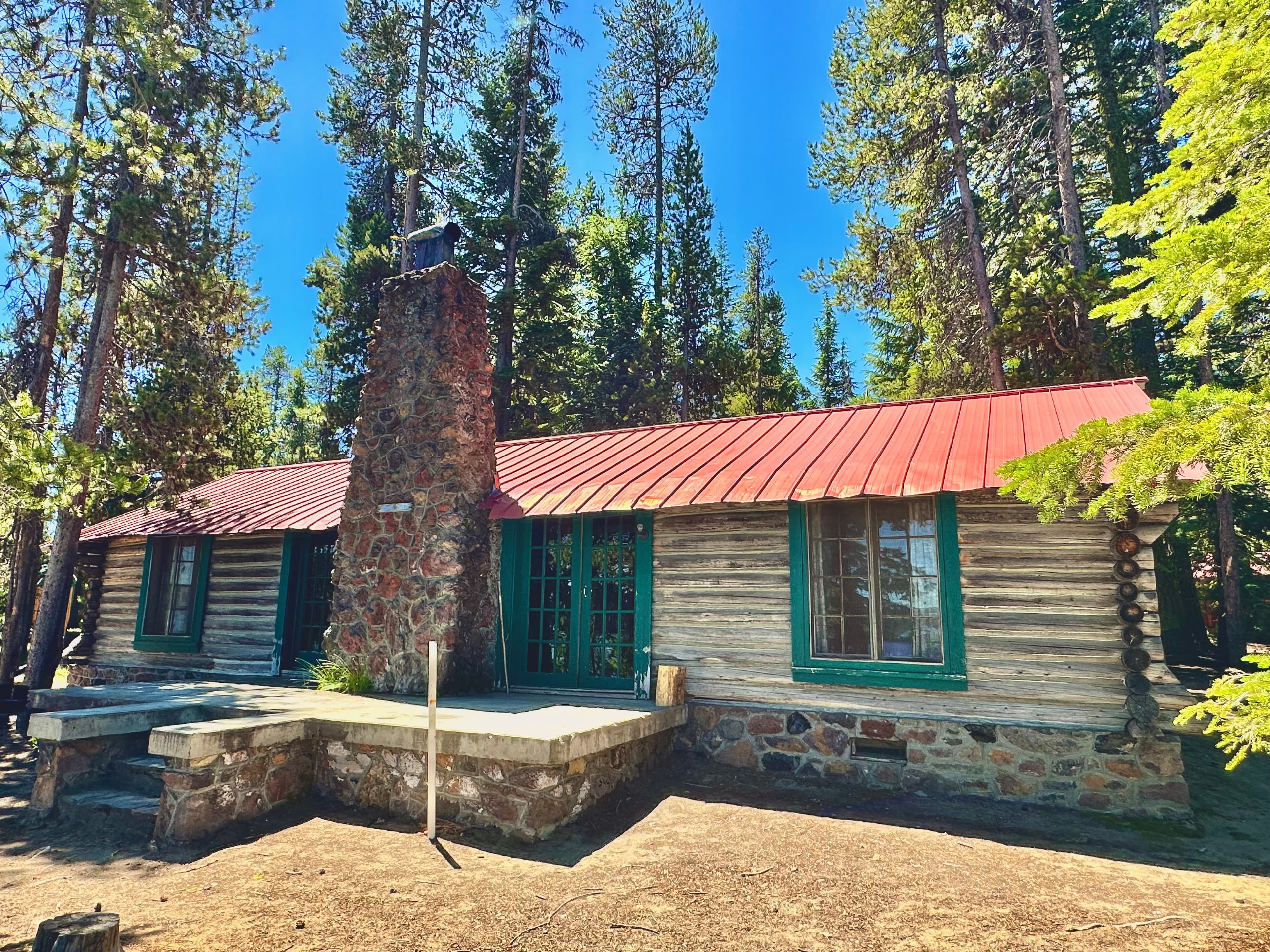 A rustic log cabin with a red metal roof, situated among tall pine trees.
