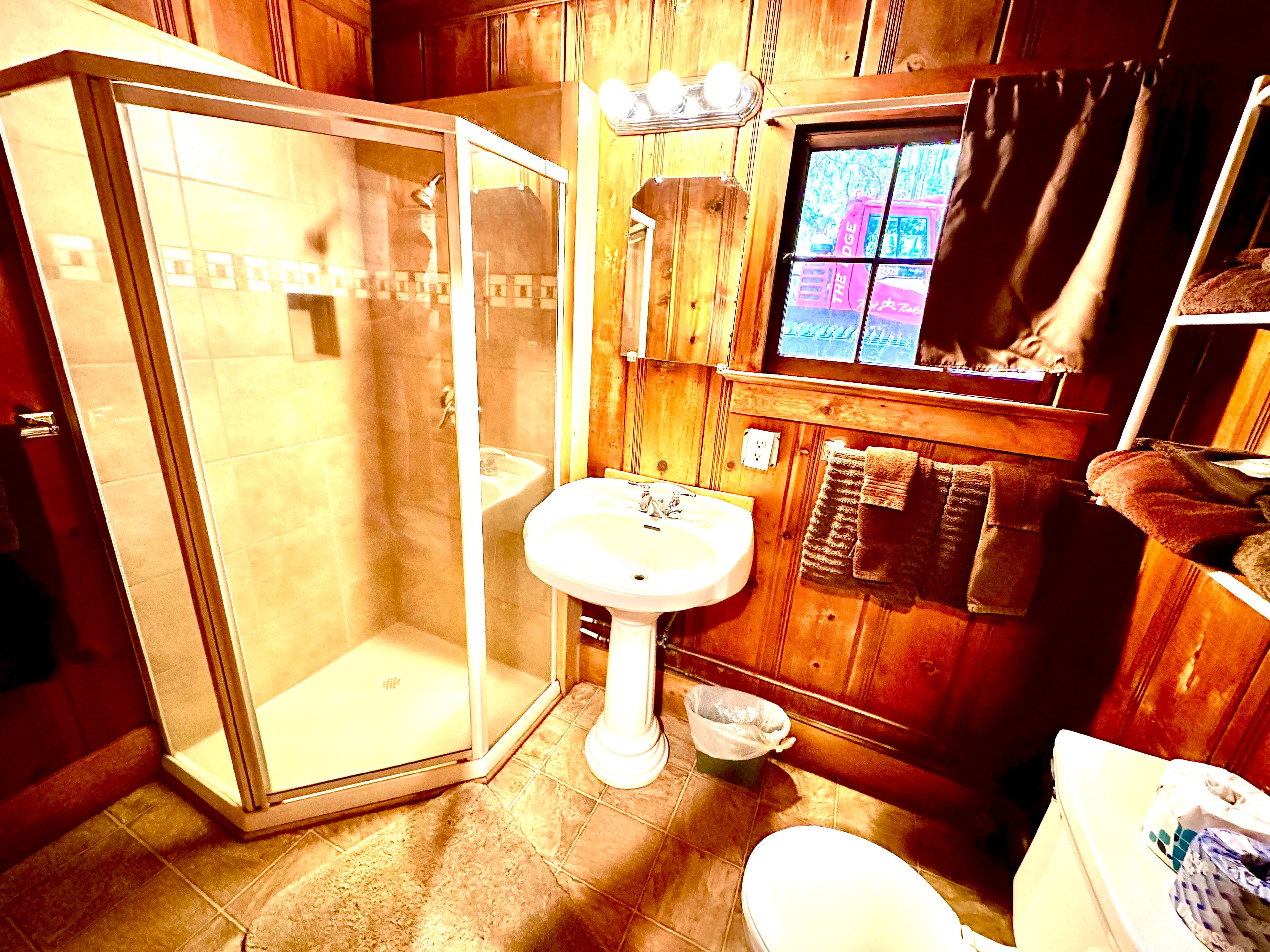 A wooden-paneled bathroom featuring a shower stall, pedestal sink, and window with curtains.
