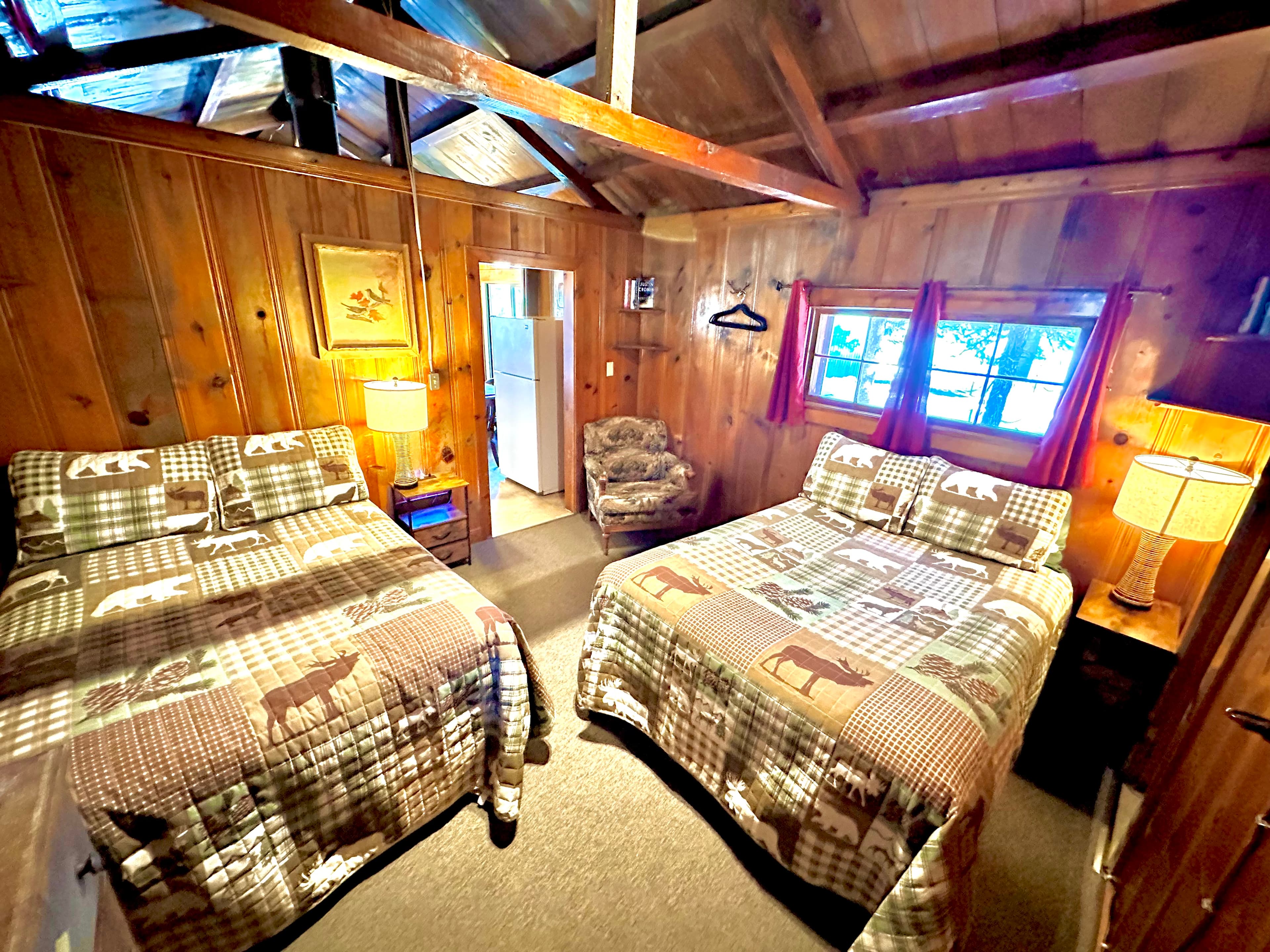 A cozy wooden cabin bedroom featuring two beds adorned with nature-themed quilts and warm lighting.