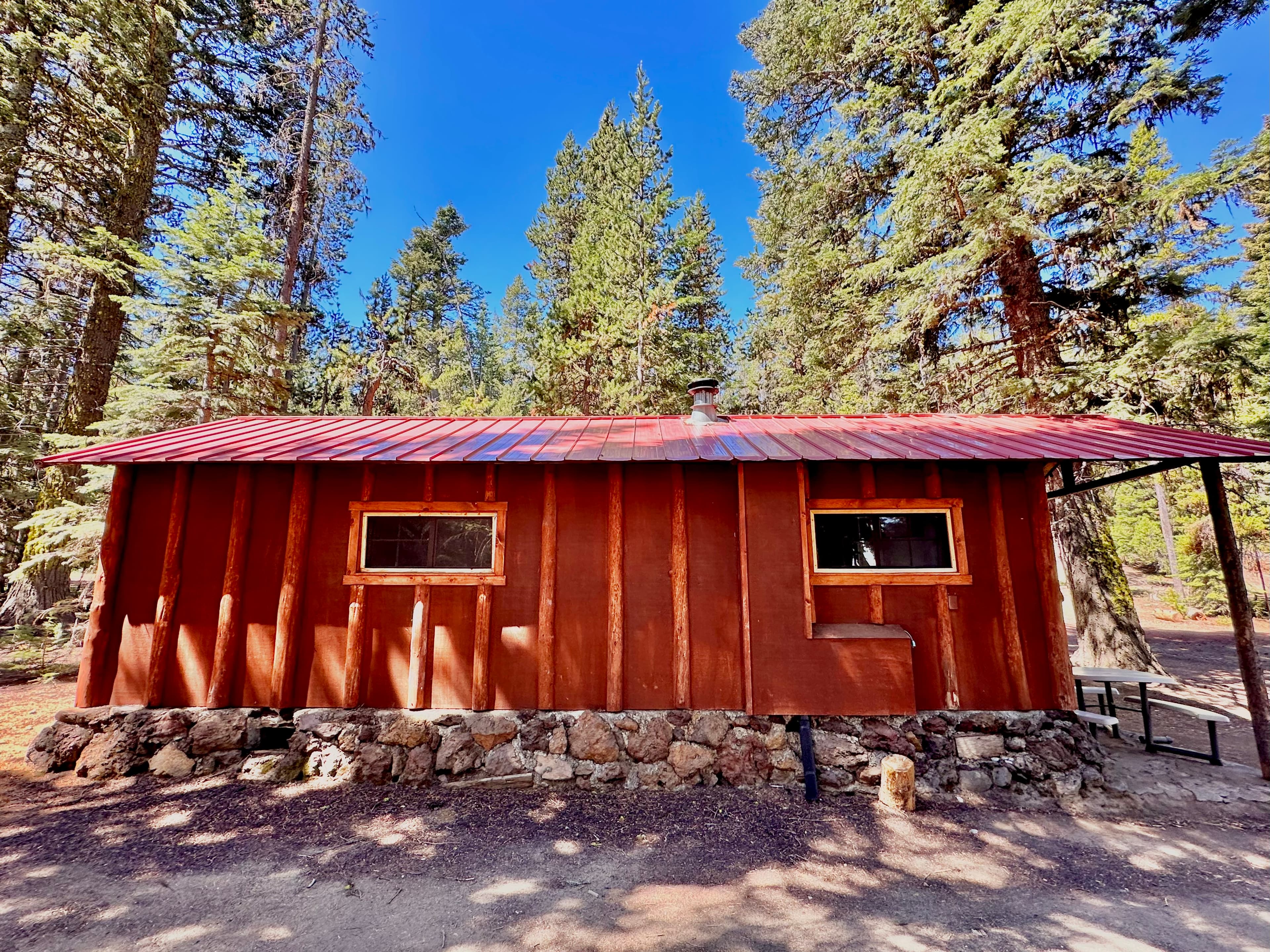 A rustic brown cabin with a red metal roof surrounded by tall trees.