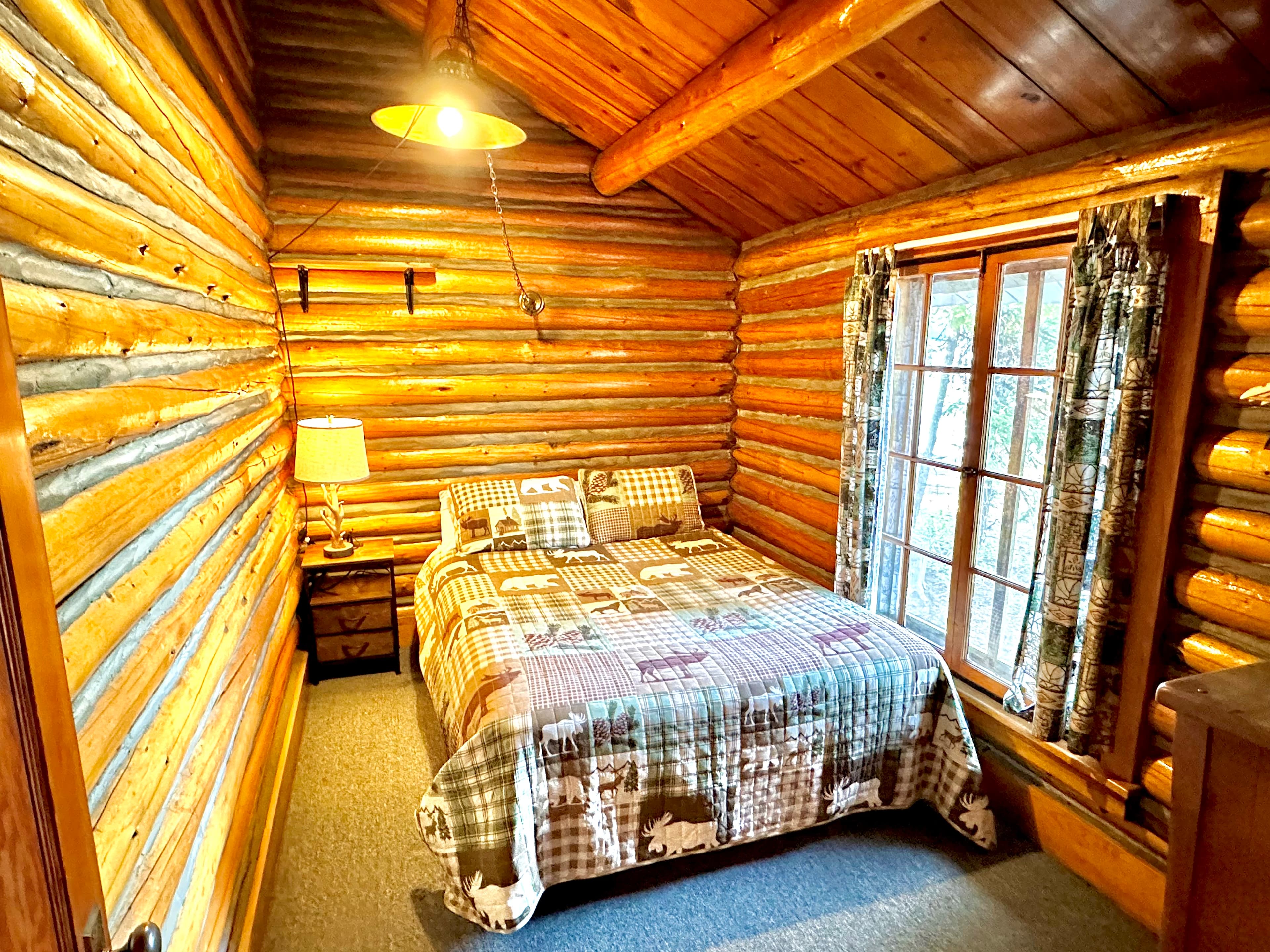 Cozy log cabin bedroom featuring a double bed with a patterned quilt, a nightstand, and a window with soft curtains.