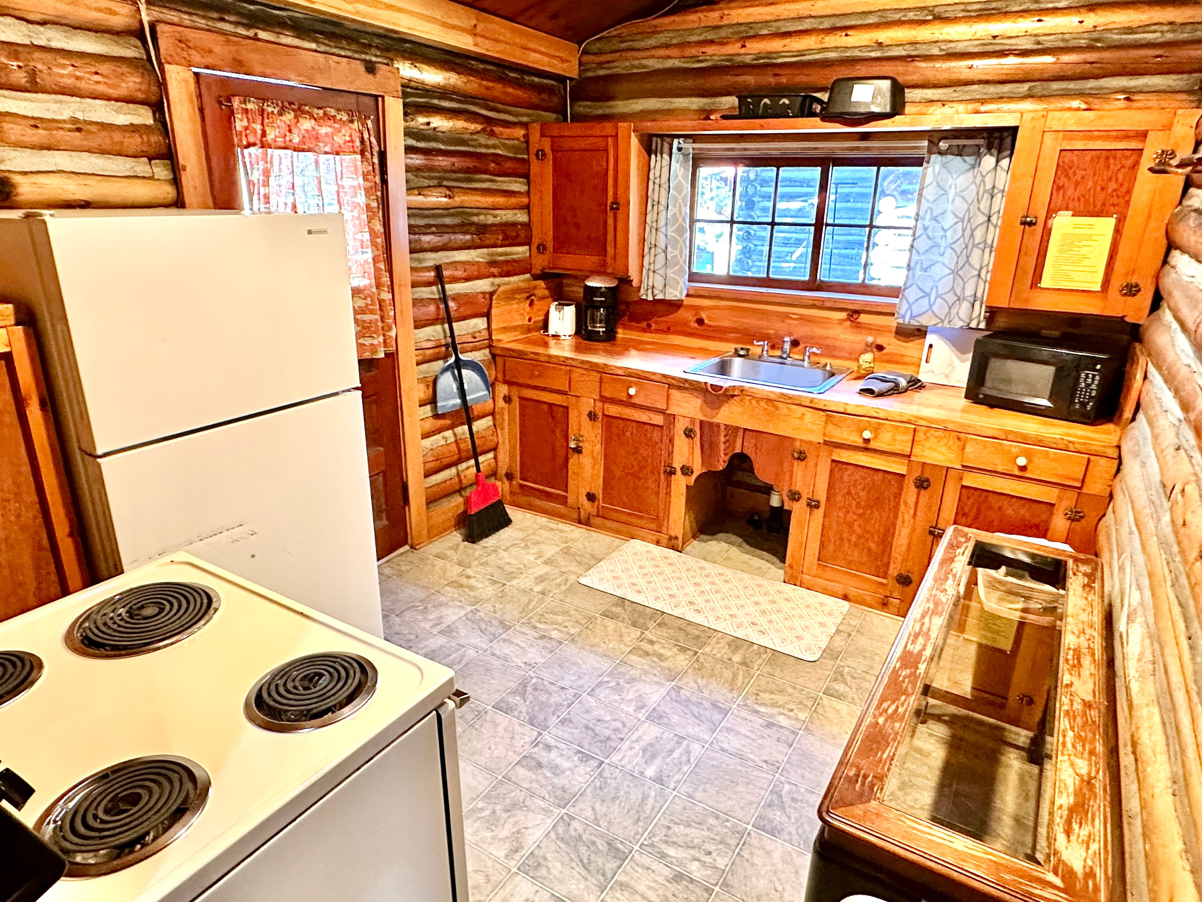 A cozy log cabin kitchen featuring wooden cabinets, a refrigerator, a stove, and a sink with a window view.