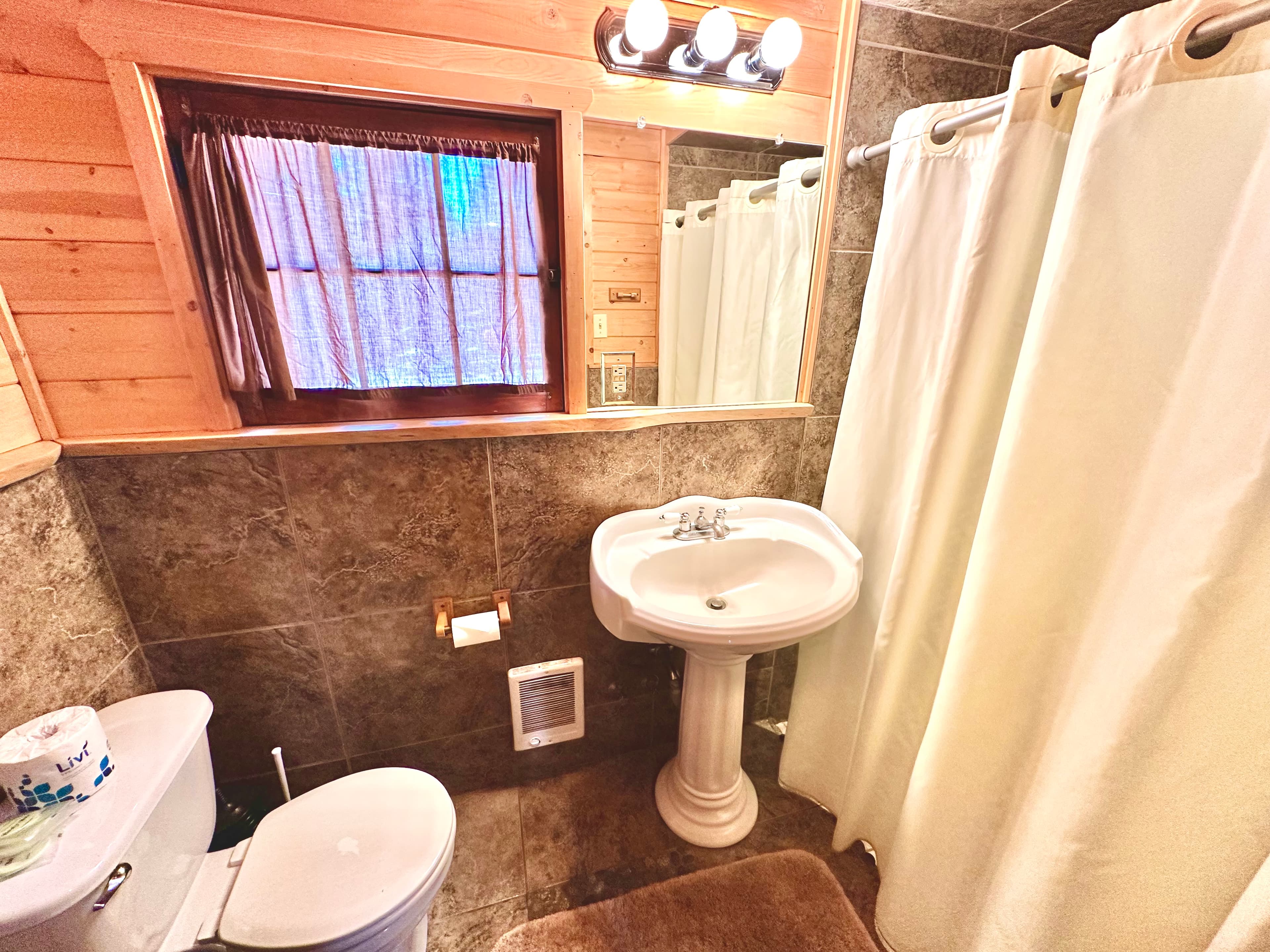 A cozy bathroom featuring a white sink, toilet, shower with curtains, and a wooden window.