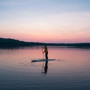 A person paddleboards on calm water during sunset.