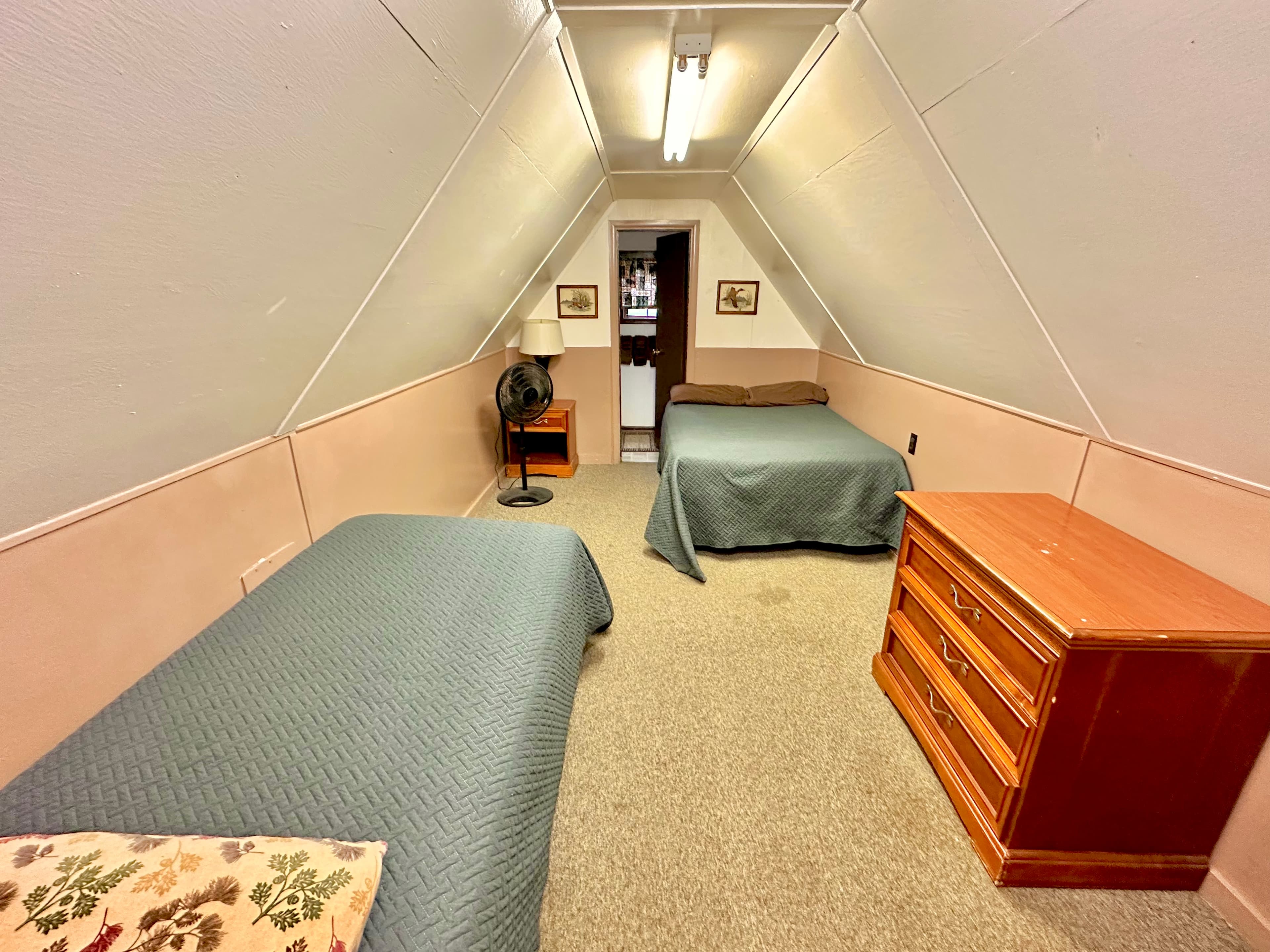 A cozy attic room featuring two beds, a dresser, and a fan under a slanted ceiling.