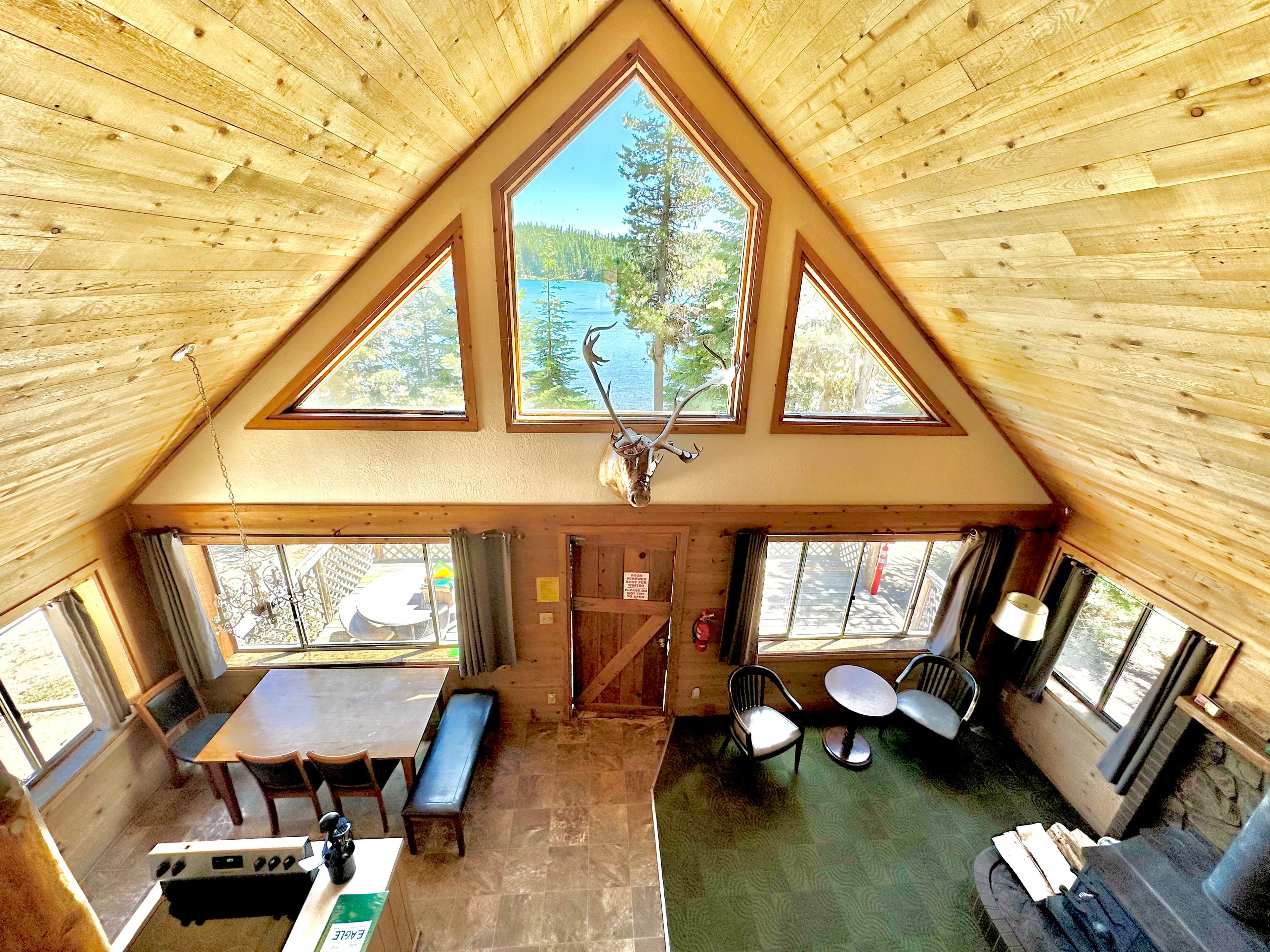 A cozy wooden cabin interior with large triangular windows overlooking a lake and forest.