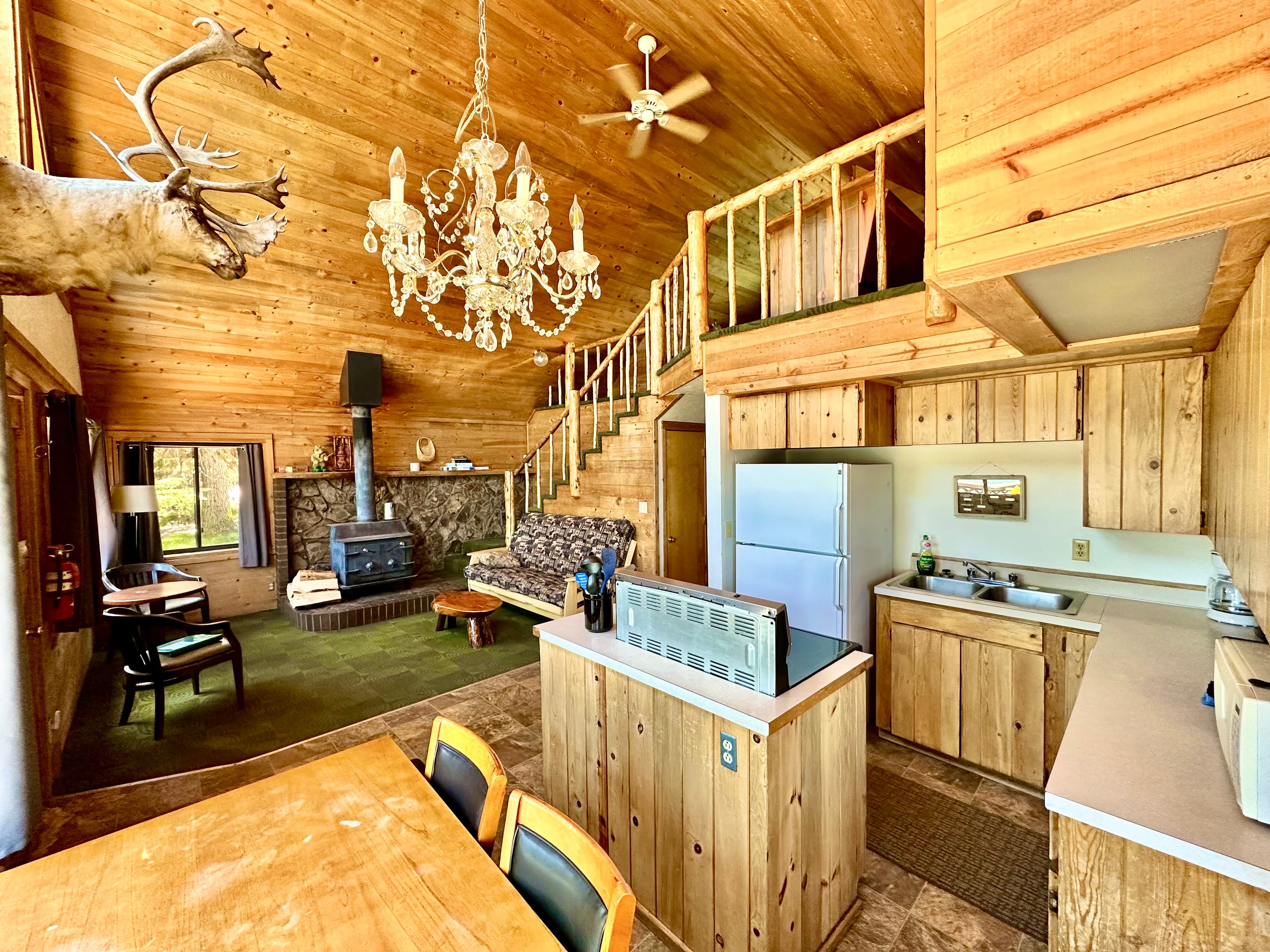 A cozy wooden interior of a cabin featuring a kitchen, dining area, and a staircase, with a chandelier and a mounted deer head adding character.