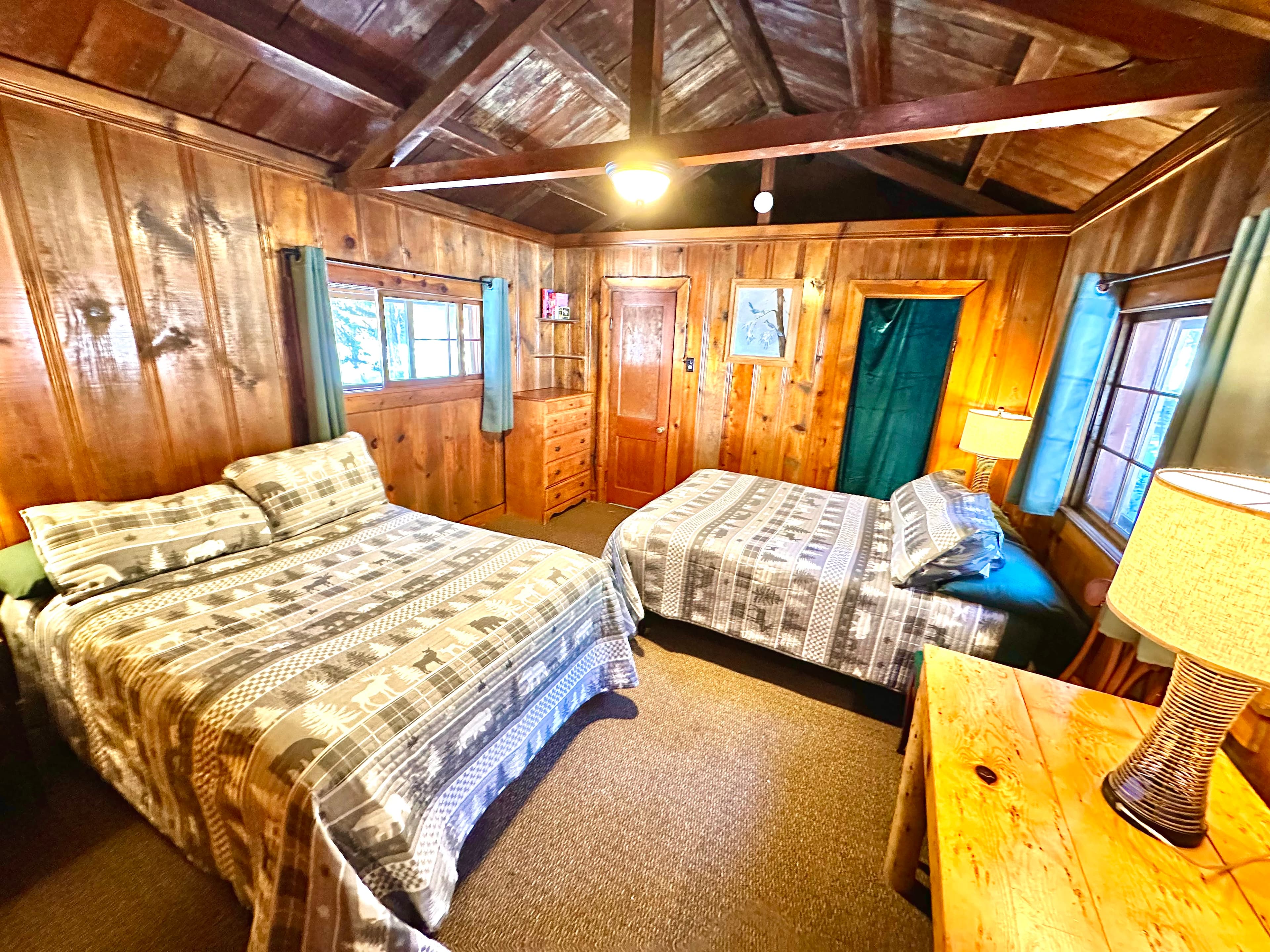 A cozy wooden cabin bedroom featuring two beds with patterned blankets and natural light coming through the windows.