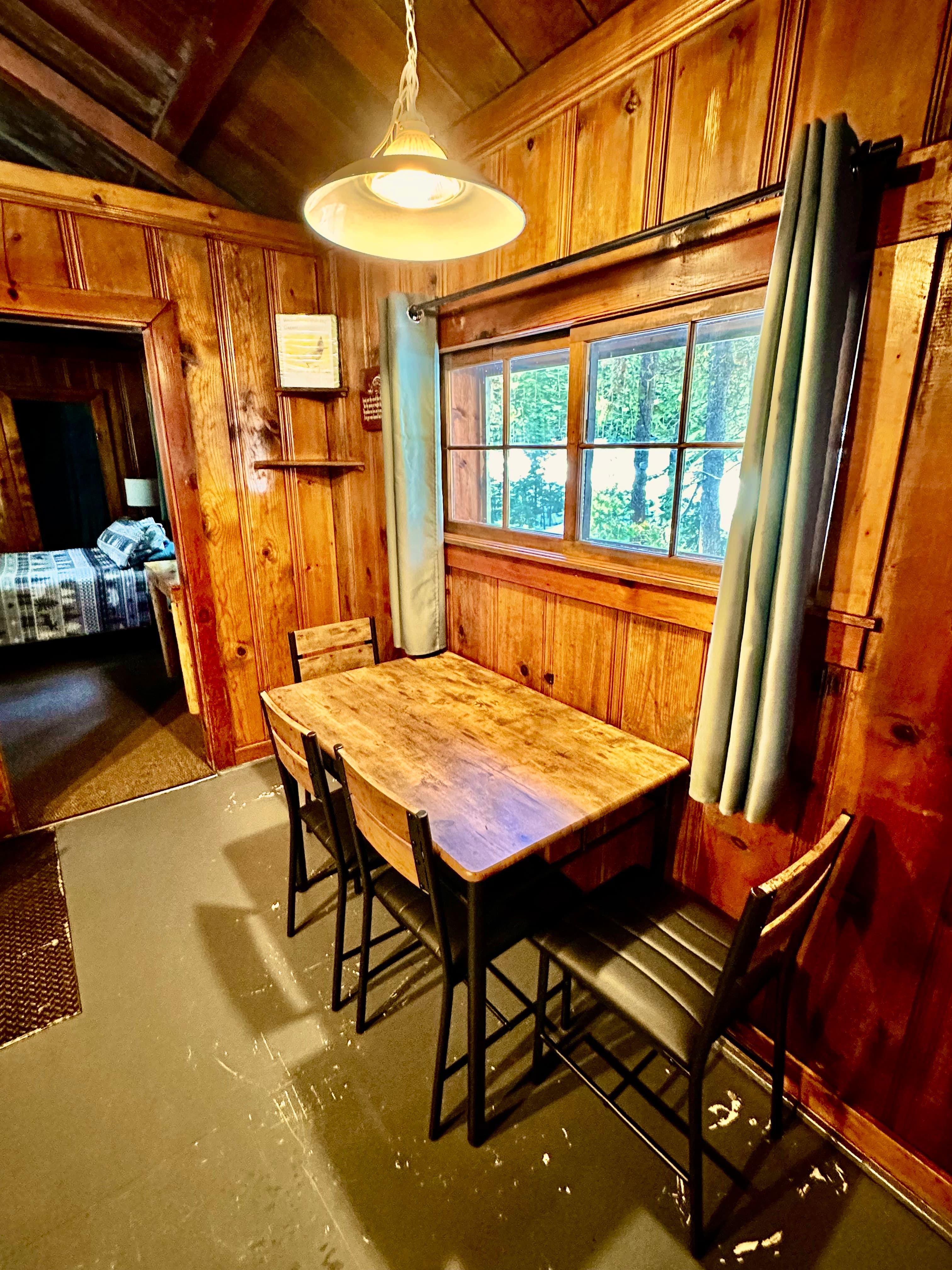 A cozy wooden dining area featuring a table and chairs, adjacent to a softly lit room with a window.