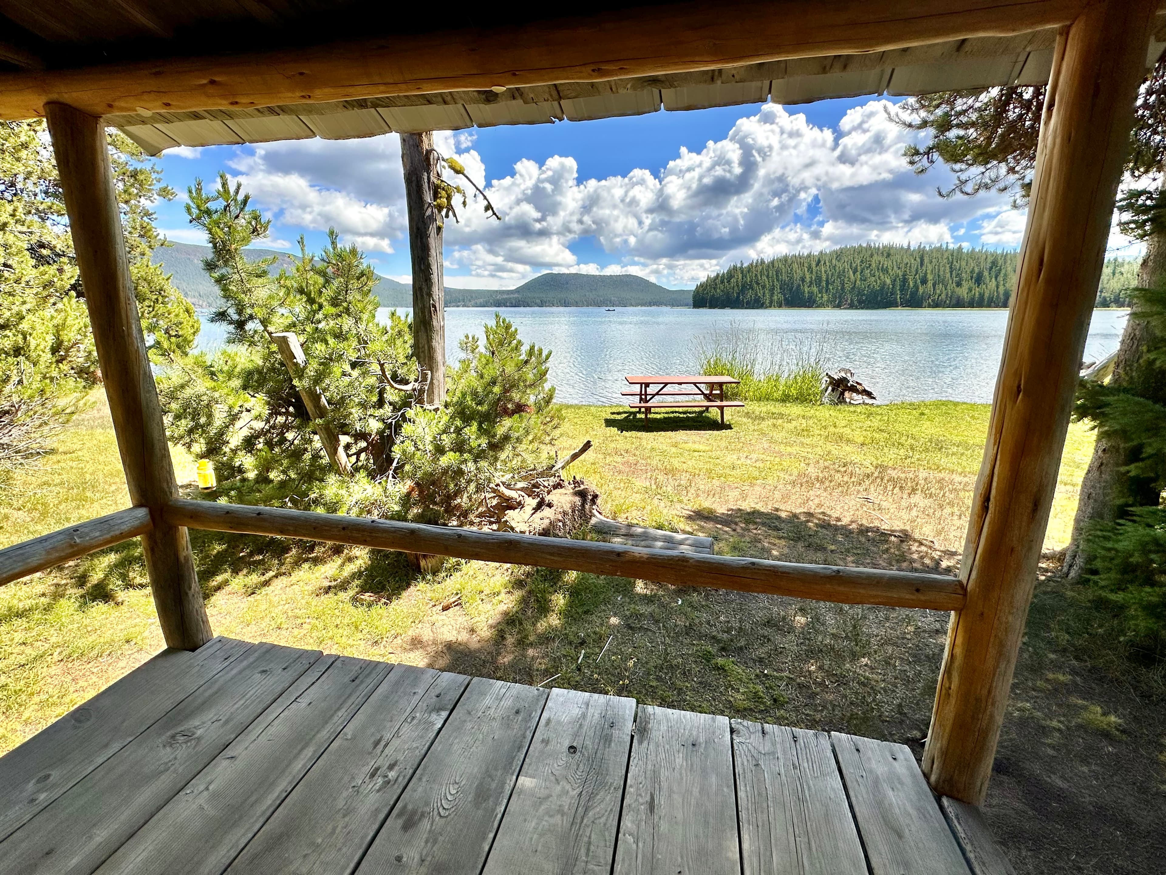 View from a wooden porch overlooking a tranquil lake with a picnic area and mountains in the background.
