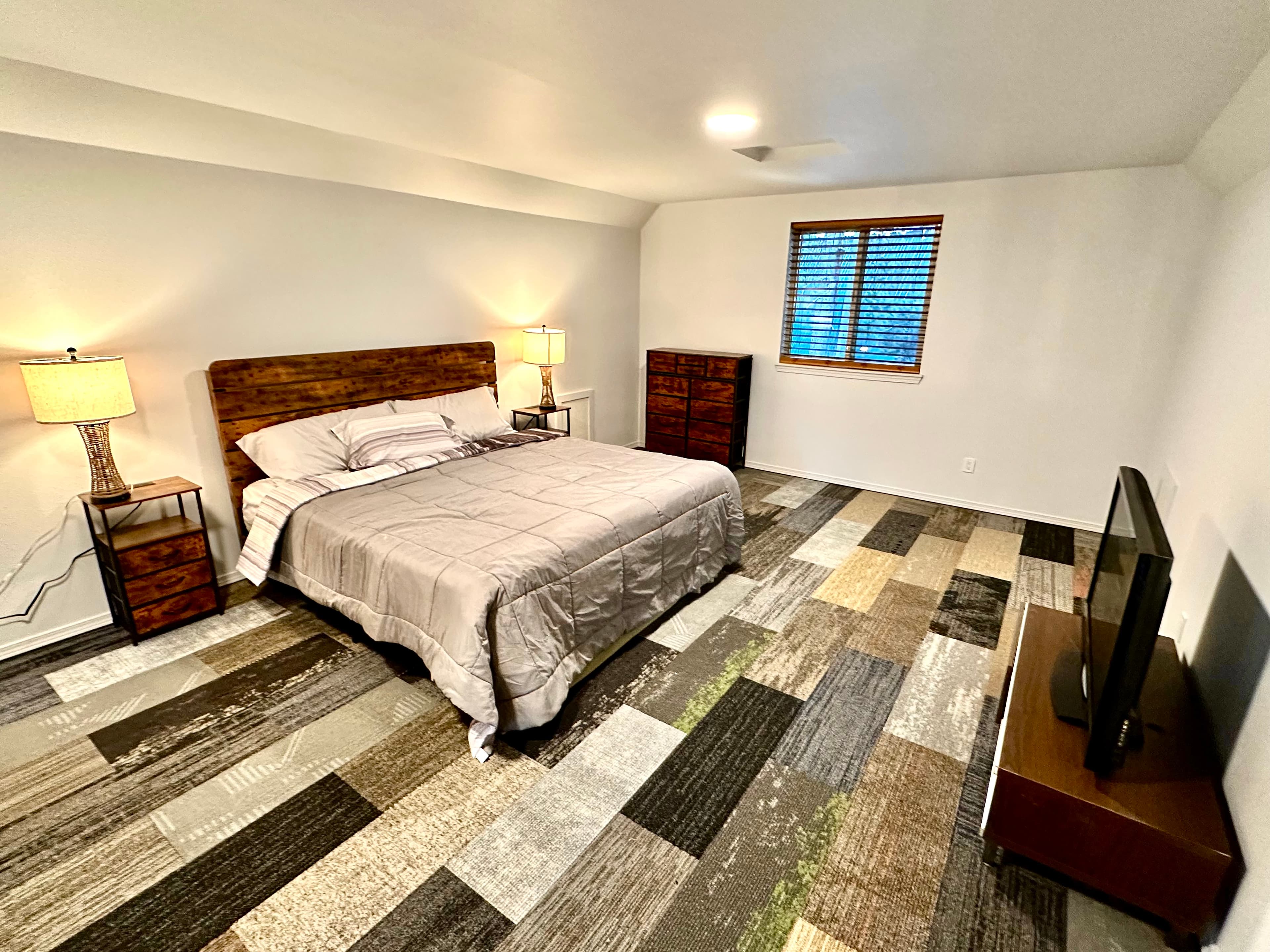 A neatly arranged bedroom featuring a queen-sized bed, two lamps, a dresser, a window with blinds, and a television.