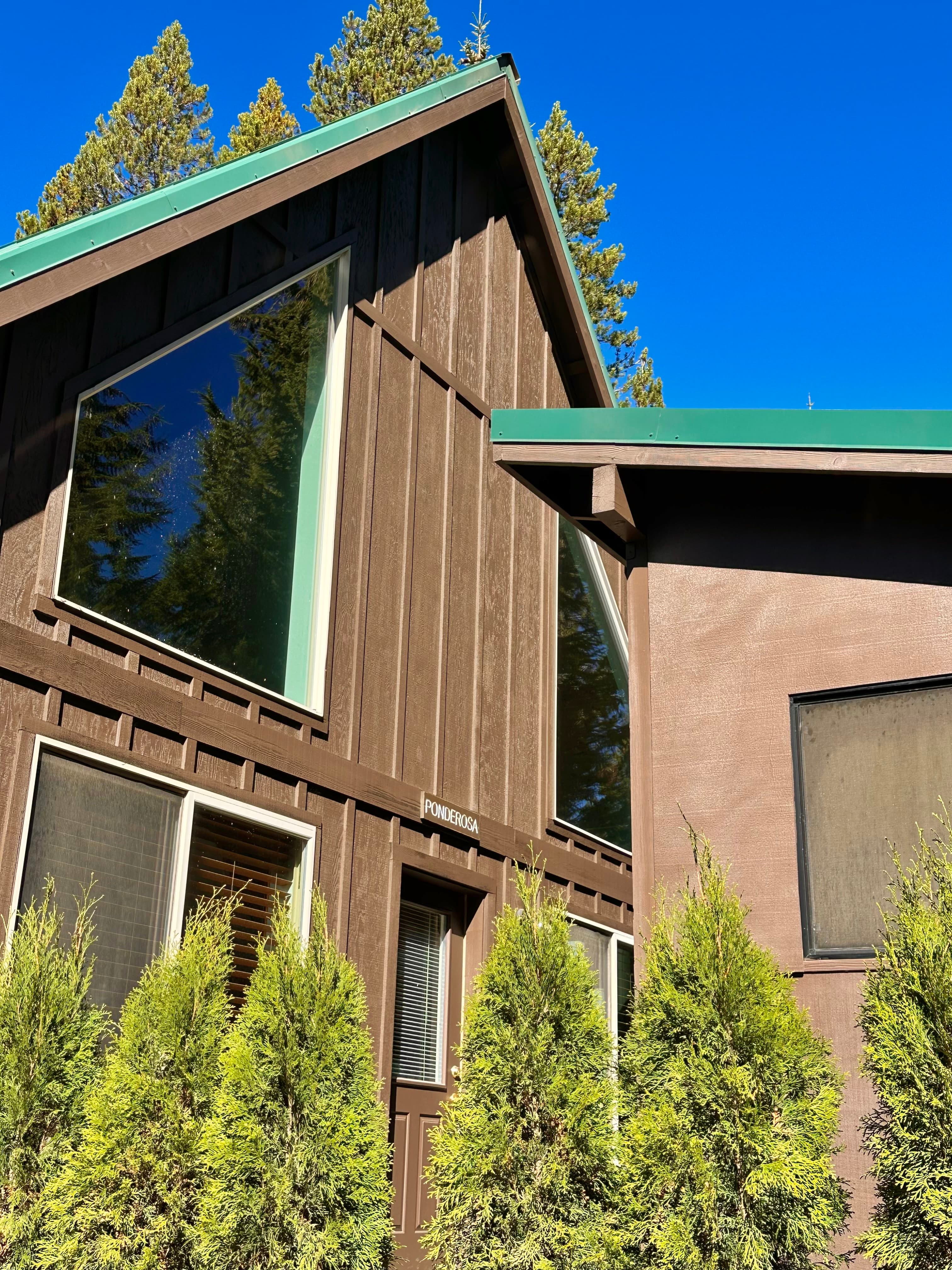 Exterior view of a brown chalet with large windows, surrounded by green shrubs against a clear blue sky.