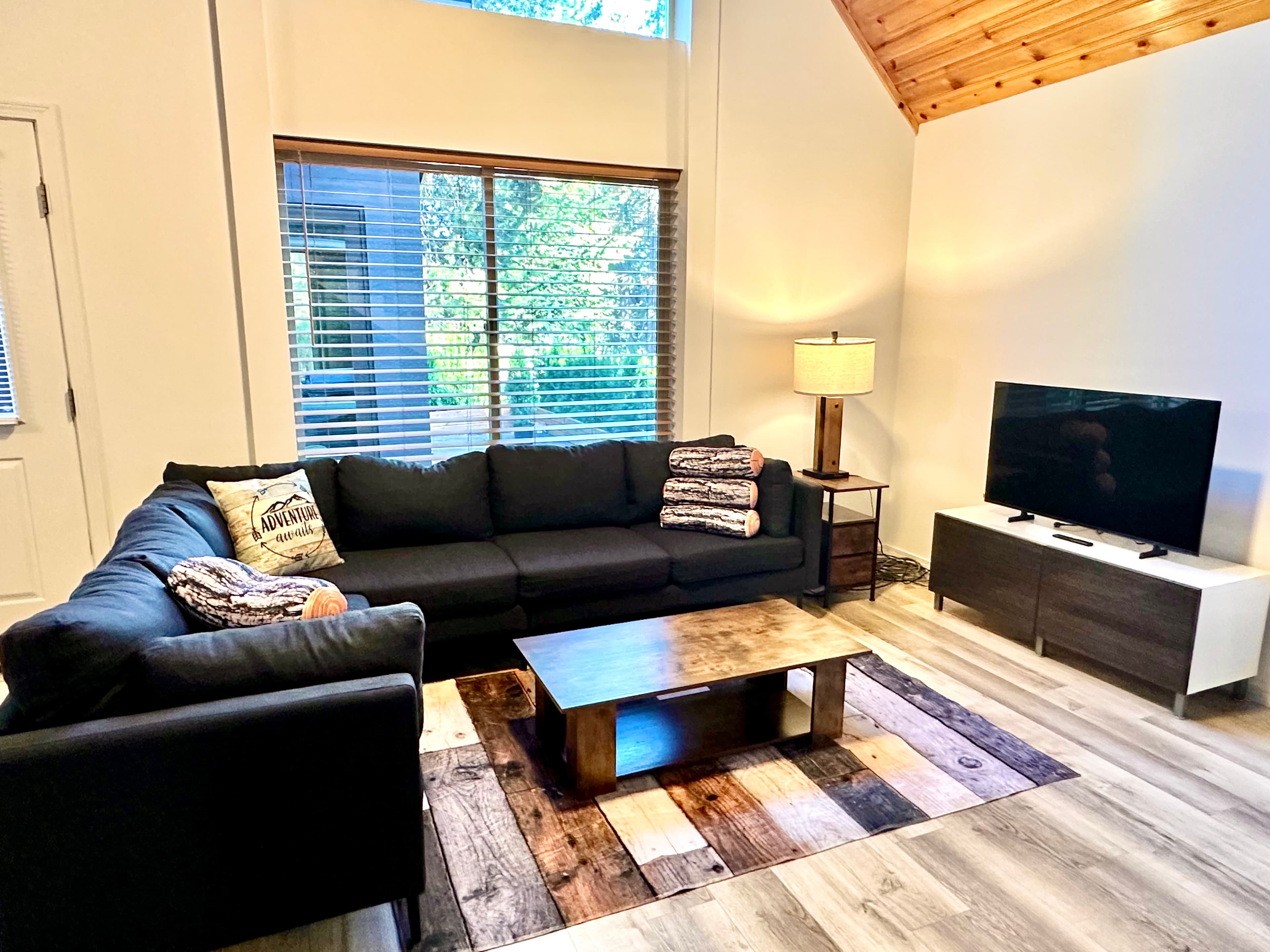 A cozy living room featuring a black sectional sofa, a wooden coffee table, a lamp, and a television against a backdrop of large windows.