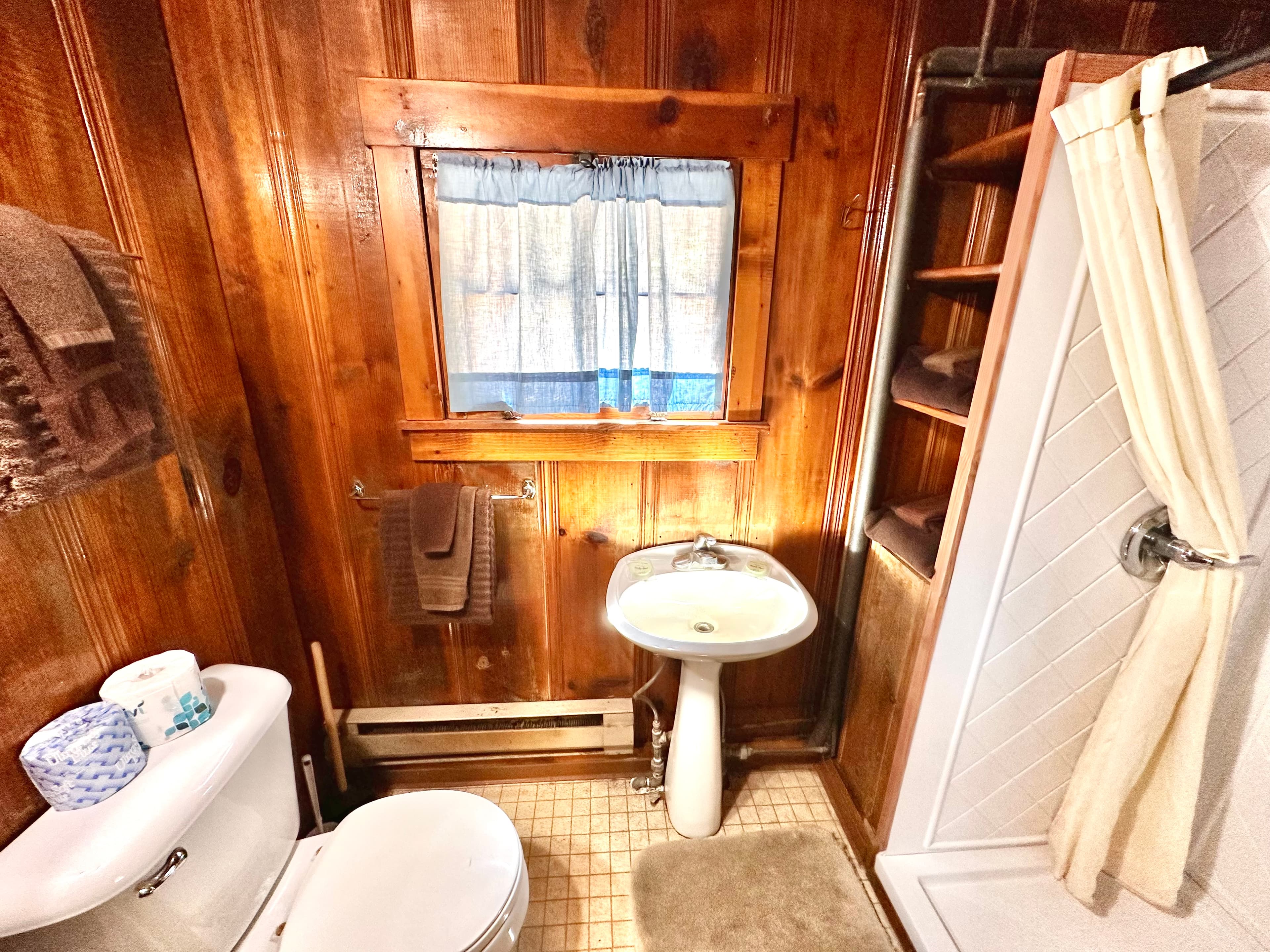 A rustic bathroom featuring wooden walls, a window with a curtain, a toilet, a sink, and a shower stall.