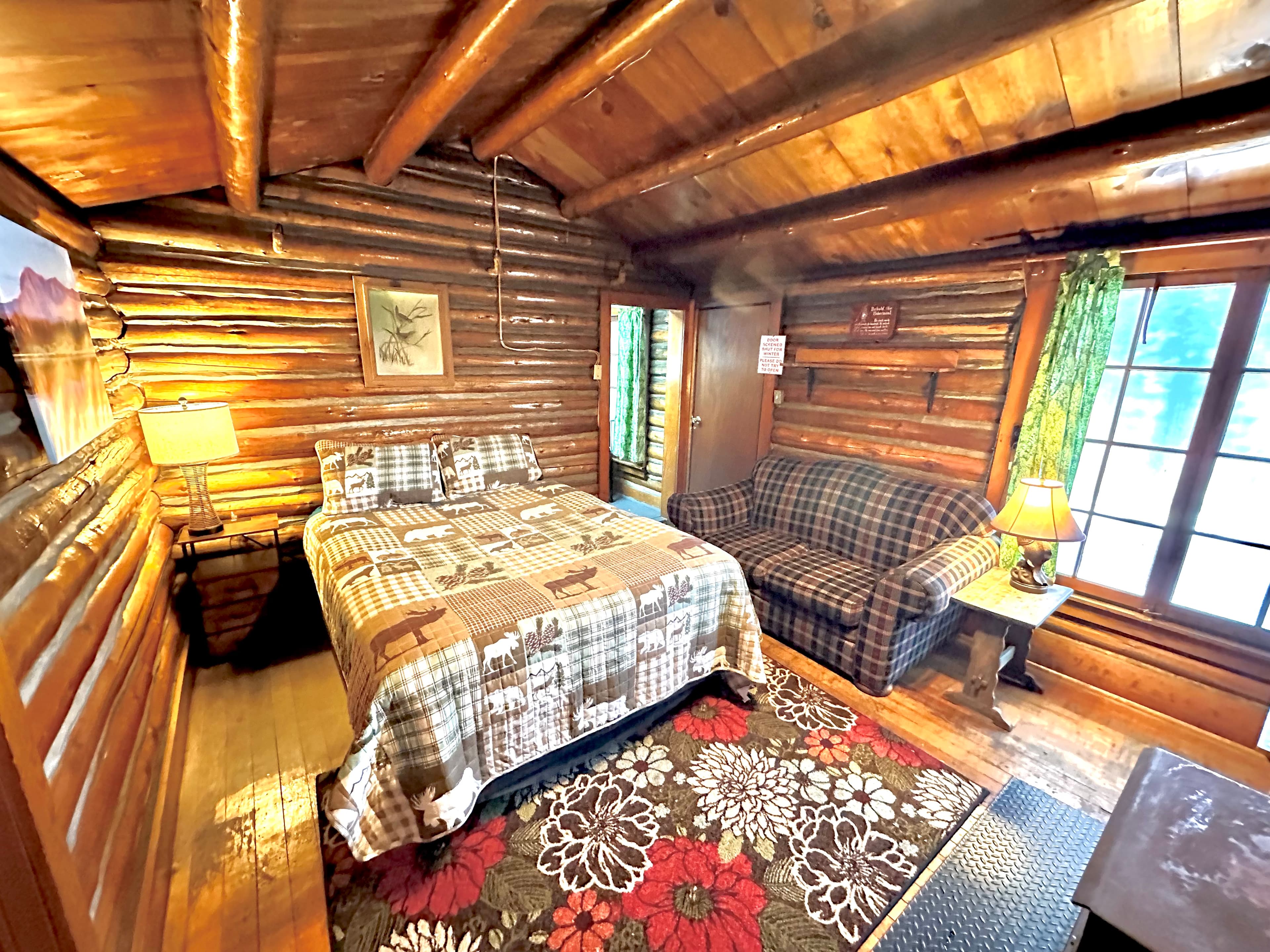 A cozy log cabin interior featuring a bed with a patterned blanket, a plaid sofa, and wooden accents.
